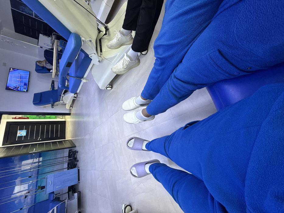 This image depicts the interior of a medical facility, possibly an infusion or dialysis center, located in Meknes, Morocco. The scene is captured from a low, first-person perspective, showcasing the extended legs of two individuals in the foreground: one on the left wears blue fleece pants and light purple slides over white socks, while the other on the right wears blue fleece pants and white slip-on shoes over white socks. Immediately to the right, a third person's legs, clad in black pants and white athletic shoes over white socks, rest in a blue and white medical recliner.
Several identical blue and white medical recliners are arranged throughout the room. One recliner in the midground is occupied by a woman wearing a hijab, partially obscured, with a small table and an IV pole positioned nearby. A wall-mounted television on the far wall displays a tennis match. The background features a dark paneled door with a blue warning sign on the left. A red fire extinguisher is mounted on the wall adjacent to the door. A large wall graphic depicting a sunny beach scene with blue water and sky covers the wall behind multiple mobile IV poles and a light blue rolling cabinet. The floor is composed of light grey tiles, and the walls are light-colored.