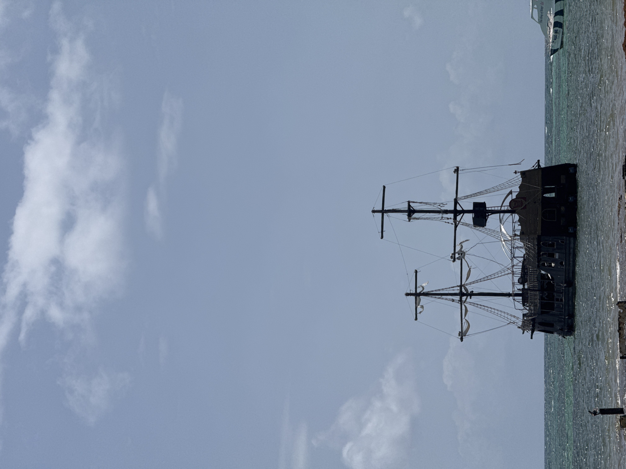 This image captures a pirate-themed ship anchored in the shallow, turquoise waters off the coast of Higüey, Dominican Republic. The ship, dark and imposing with multiple masts and tattered sails, dominates the right side of the frame. The water, with its gentle ripples and sparkling surface, suggests a sunny day. The sky is a pale blue, dotted with wispy white clouds, indicating fair weather. In the upper right corner, a modern white boat with black trim and lettering is partially visible, creating a contrast between the old and new forms of maritime vessels. The overall scene evokes a sense of adventure and a tropical atmosphere. There is a faint inscription on the modern boat that reads "The King of Kings".