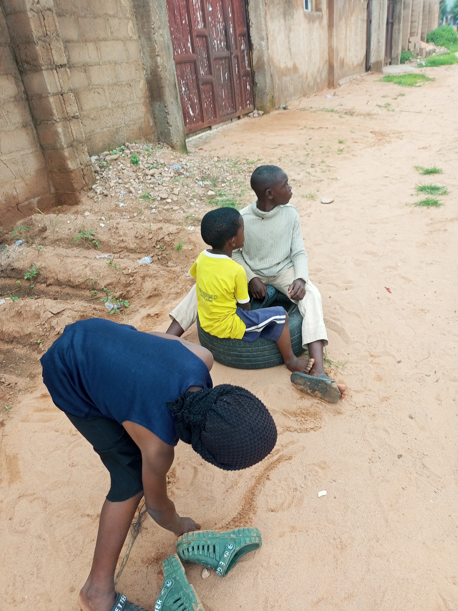 In the city of Tempure, Nigeria, three young children are interacting in a sandy outdoor area with a rough, unpaved surface. The background features a simple, aged building constructed from bricks, with a weathered red door. The ground is a mix of sand and sparse vegetation.
One child, wearing a navy blue shirt and dark pants, is bent over and appears to be digging or playing with the sand. Another child, in a bright yellow t-shirt and blue shorts, sits on a black tire. This child is looking to the right. The third child, dressed in a light-colored sweater and light-colored pants, is also sitting on the tire, positioned slightly behind and to the right of the child in yellow. This child is also looking to the right.
Several pairs of green, hole-covered sandals are visible on the ground near the child who is bending over, and one pair of dark sandals is on the foot of the child in light-colored pants. The overall scene suggests a casual moment of play or activity among children in a modest urban or semi-urban environment.