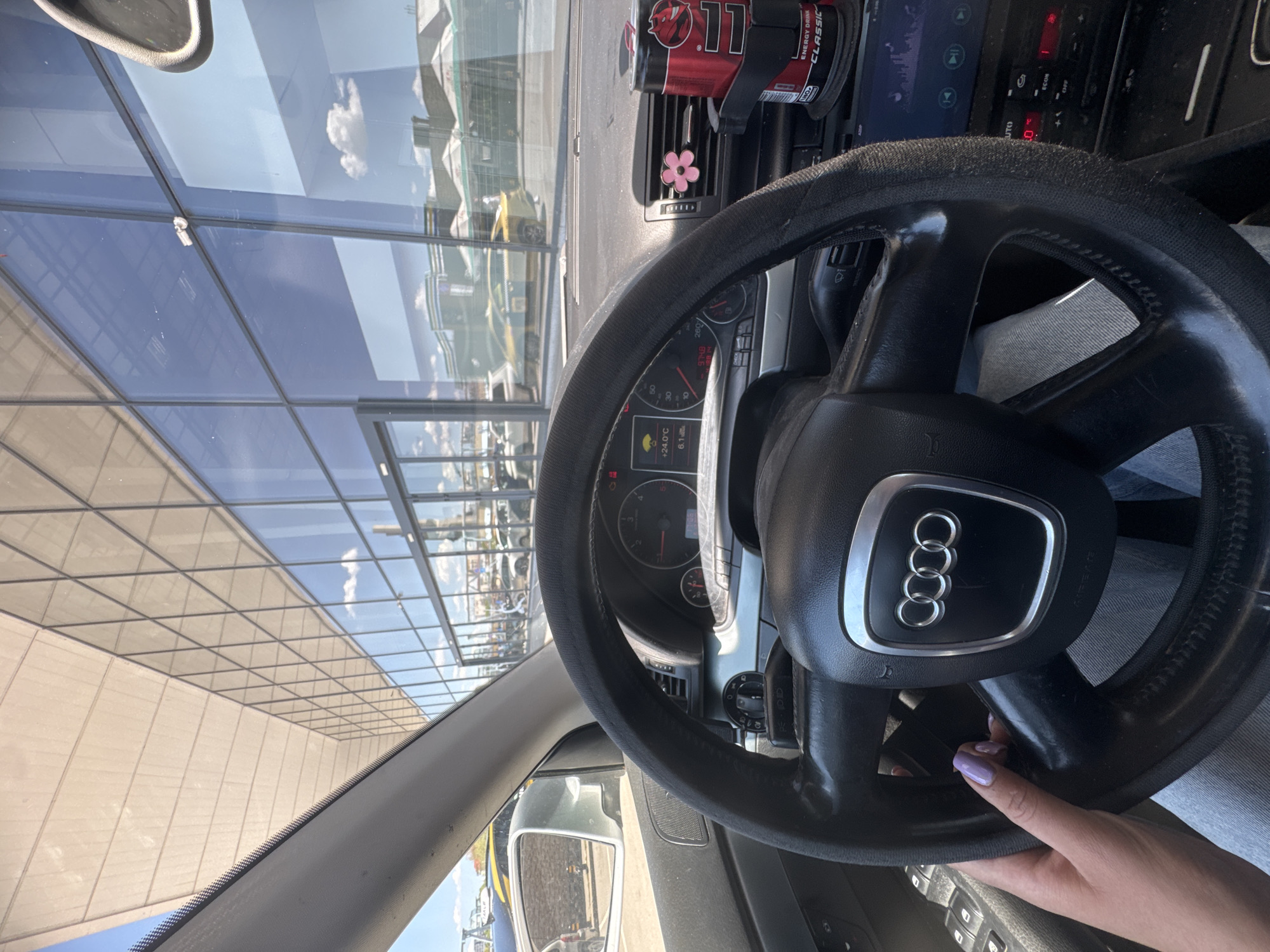 The image depicts the interior of an Audi car, specifically the steering wheel with the Audi logo prominently displayed. A person's hand, with lavender-colored fingernails, is lightly holding the steering wheel. The car appears to be parked or stationary. The dashboard is visible, showing the car's controls and a can of "Energy Drink Classic" in a holder near the air vent. The car's temperature reading indicates +24.0°C. Through the windshield, a modern building with large glass windows is visible, reflecting the sky and some clouds. The building's architecture suggests it may be a public building, possibly an airport.
Based on the architecture and reflective glass, the location might be near Sofia Airport in Bulgaria. This is an educated guess based on the modern architecture, reflective glass, and the potential for reflection of aircraft in the glass. A more precise identification of the location would require additional visual context.
