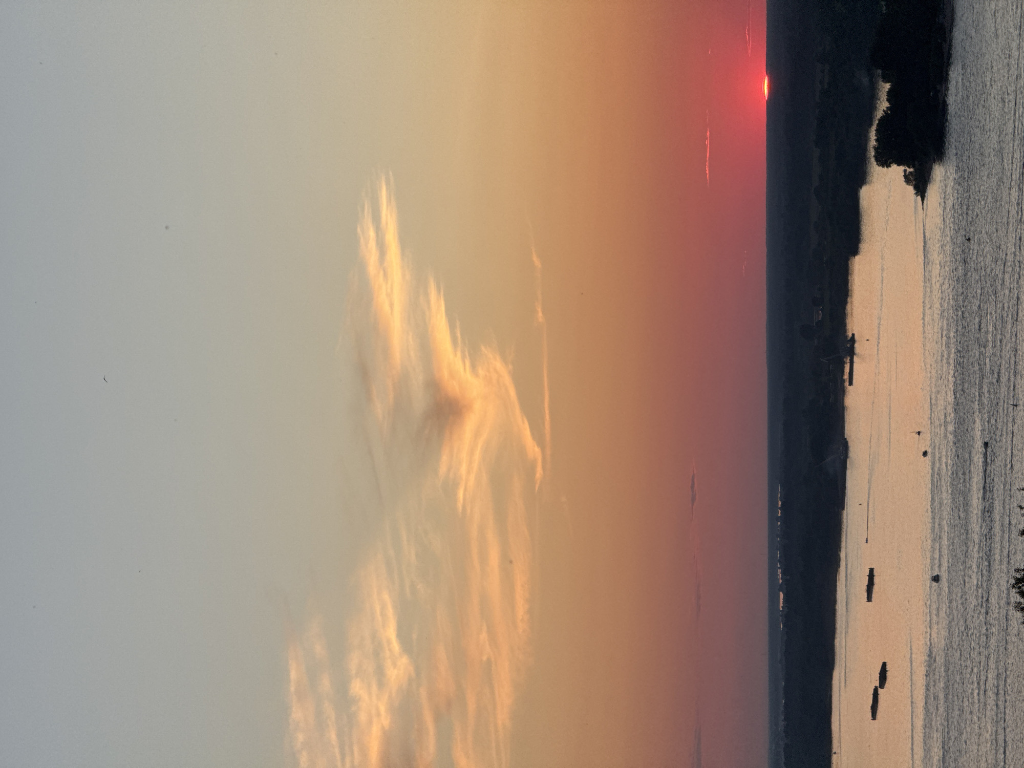 This is a photograph of the sunset over the Volga River in Nizhny Novgorod, Russia. The sky is a gradient of colors, from pale blue at the top to orange and pink near the horizon. Wispy clouds are scattered across the sky, catching the sunset light. The river is wide and calm, reflecting the colors of the sky. Several barges and boats are visible on the water. The far bank of the river is lined with dark trees, and the silhouette of a city skyline can be seen in the distance. The sun is a bright red orb, just touching the horizon.