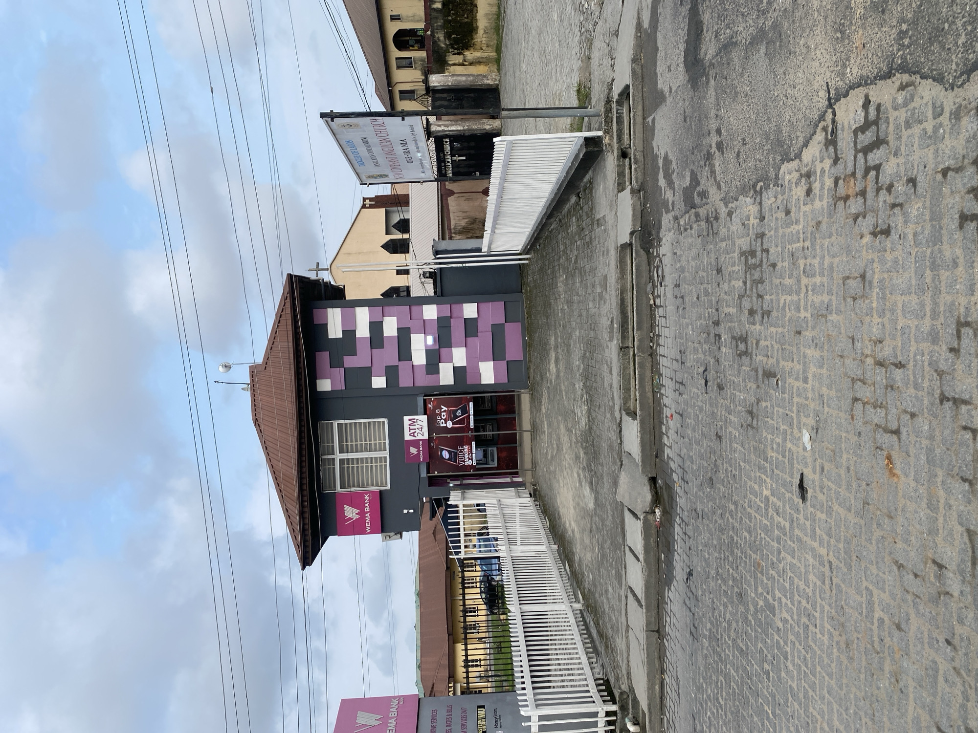 The image shows a Wema Bank branch located in a city, possibly in Nigeria, given the signage. The building has a distinctive facade with purple and grey geometric patterns. An ATM is visible outside the bank, along with signs for "Voice Banking" and "Tap & Pay". A white picket fence encloses the bank's premises.
To the right of the bank, a sign indicates "DIOCESE OF LAGOS ANGLICAN COMMUNION NEW COVENANT ANGLICAN CHURCH OXE-IRA NLA". This suggests the presence of a church nearby, and the building with a cross visible in the background confirms this. The street in front of the bank is paved with interlocking bricks. The sky is overcast with scattered clouds. The overall scene depicts an urban environment with a mix of commercial and religious institutions.