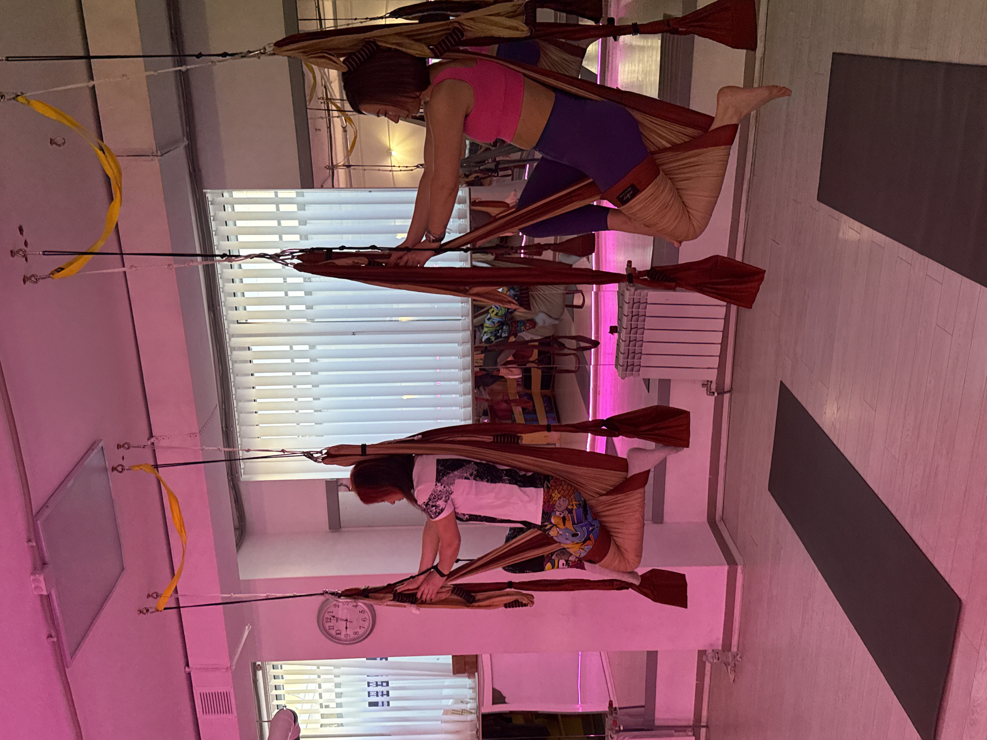 Two women are engaged in an aerial yoga or fitness session within a brightly lit studio in Chaikovsky, Russia. They are suspended in individual fabric hammocks or slings that hang from the ceiling.
The woman on the right wears a pink sports bra and purple leggings, with bare feet. She is positioned with her torso slightly forward, hands resting on the fabric, and her legs wrapped within the beige part of the sling, demonstrating a focused posture. The brand name "Loop" is visible on the beige fabric of her hammock.
The woman on the left is dressed in a white t-shirt with a dark pattern and patterned shorts, wearing white socks. She holds a similar pose, supported by her own hammock, which appears to be a combination of reddish-brown and beige fabric.
The studio features a light wooden floor, and a dark gray yoga mat is laid out on the right side. White horizontal blinds cover windows along the back wall, suggesting natural light, although the room is illuminated by artificial white and vibrant pink lighting, creating a dynamic atmosphere. Overhead, multiple suspension points are visible on the white ceiling, from which the hammocks and a yellow fabric loop are attached with black cords and metal fixtures. A white radiator is present near the back wall. On the left wall, a round analog clock shows the time around 10:15 or 10:20. Both women appear concentrated on their exercise, indicating a class or training session.