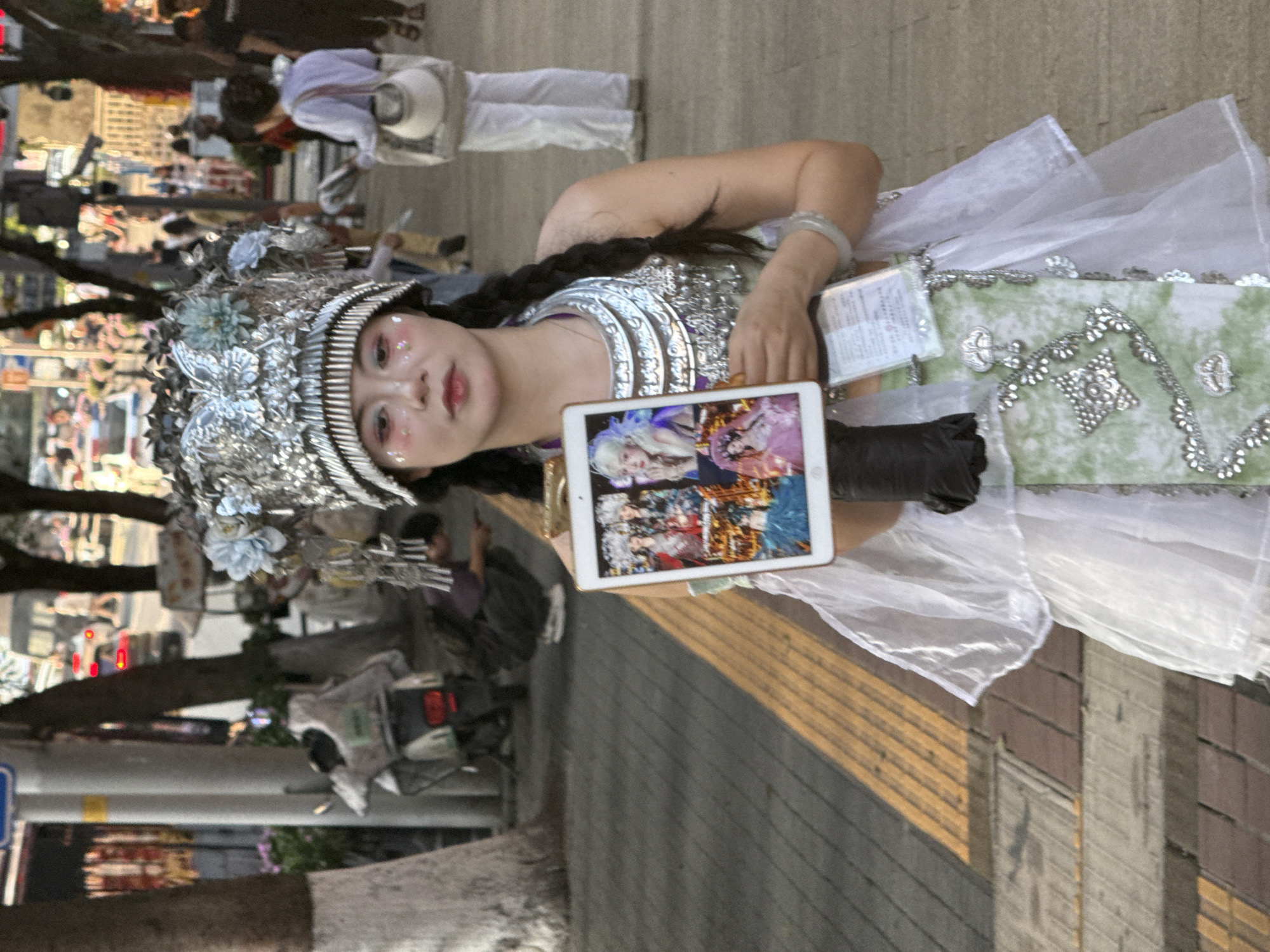 A captivating full-shot image features a person in an elaborate, decorative costume standing on a paved sidewalk, likely in Yuzhong District, China. The central figure wears a striking silver headdress adorned with intricate patterns and light blue floral accents. Their face is decorated with artistic makeup, including glitter around the eyes and pink-tinted lips, and they look directly at the camera with a composed expression.
The costume includes a silvery, embellished top and a light green and white skirt-like garment, also decorated with silver patterns and a sheer white overlay. A long, dark braid is visible, and a light-colored bracelet adorns their left wrist. In their left hand, they hold a white tablet (possibly an iPad) displaying several vibrant images of other elaborately costumed figures, suggesting a connection to their own attire or performance. A small white card, possibly an identification or informational card with unreadable Chinese characters, is visible tucked into their costume.
The background reveals a bustling urban environment, though blurred, with other pedestrians and trees lining what appears to be a street or public walkway. The ground is paved with distinct yellow markings. The lighting suggests it is daytime, possibly late afternoon, creating a soft glow on the subject and their surroundings. The person appears to be showcasing their elaborate outfit, possibly as a street performer, artist, or participant in a cultural event.