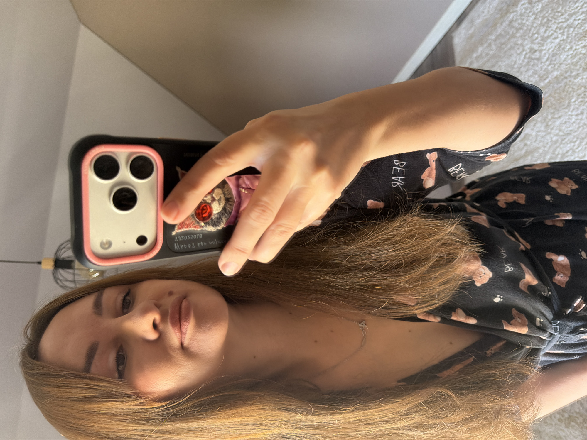 A young woman with long, auburn hair is taking a selfie in what appears to be a bedroom. She is wearing a black V-neck top adorned with a pattern of small teddy bears. A silver necklace with an 'A' pendant is visible around her neck. Her left hand is holding up a smartphone, which has a distinctive black case featuring a cartoon cat with red eyes. The background shows a sloped ceiling, part of a light fixture, and a wall. The room is illuminated by natural light, casting soft shadows.
