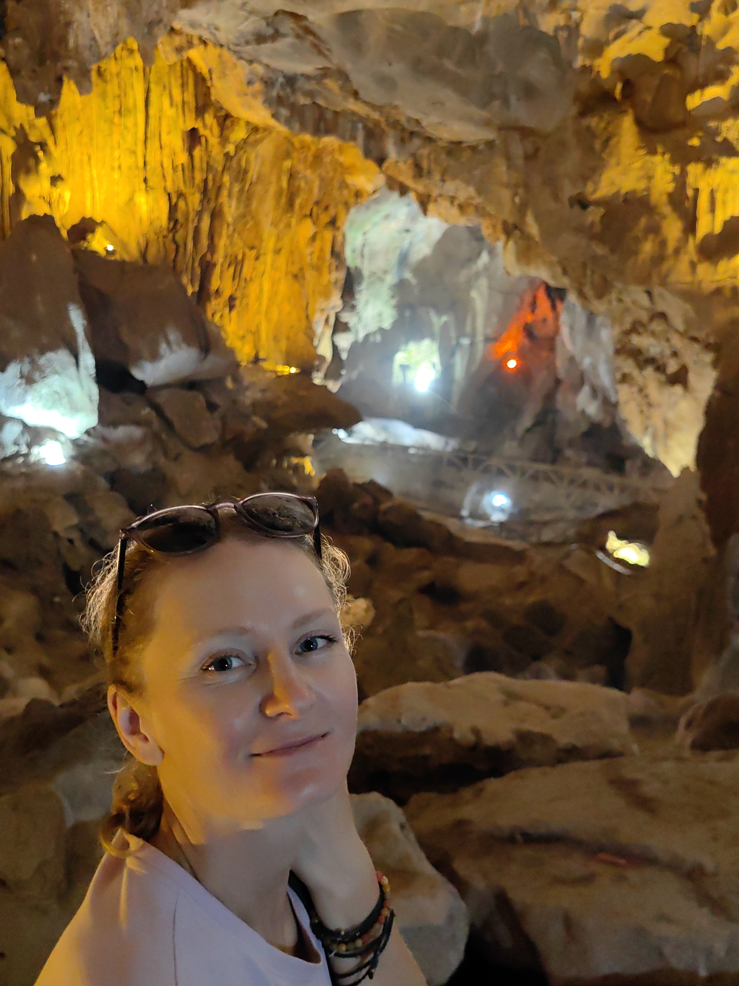 A woman with fair skin, wearing sunglasses atop her head and a bracelet, is posing for a selfie in a cave. The cave's interior is illuminated with orange and white lighting, highlighting rock formations and a walkway in the background. The location is within a cave complex located in the city of Lạng Sơn, Vietnam.