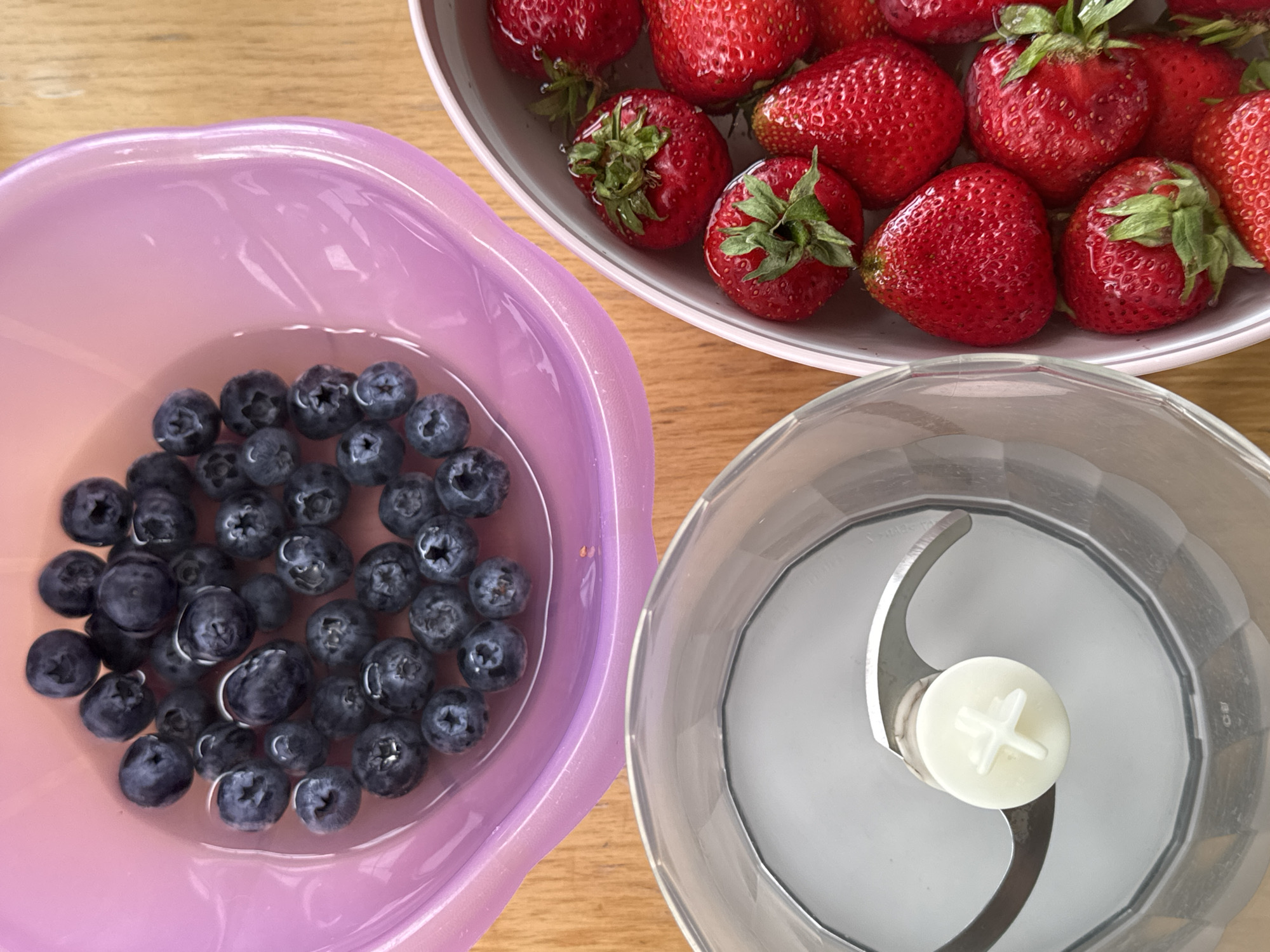 The media file shows a top-down, close-up shot of three bowls on a light brown wooden surface, likely a table.
In the foreground to the left is a large, light purple bowl filled with fresh blueberries submerged in water. The blueberries are a deep blue-black color, and many are partially submerged, reflecting the light.
To the right of the purple bowl, and slightly further back, is a clear blender cup containing a silver blade assembly with a small white piece at its center. The blender cup is empty, ready for use.
In the upper right quadrant of the image is a light grey or white bowl filled with ripe strawberries. The strawberries are a vibrant red, with green stems and leaves. Some of the strawberries are partially submerged in water within the bowl, similar to the blueberries.
The overall scene suggests preparation for making a smoothie or other blended fruit dish. The lighting appears bright and natural, indicating it could be daytime. There are no people visible in the image, and no discernible text. The setting appears to be a kitchen or dining area. The presence of fresh fruit and a blender suggests a domestic context.