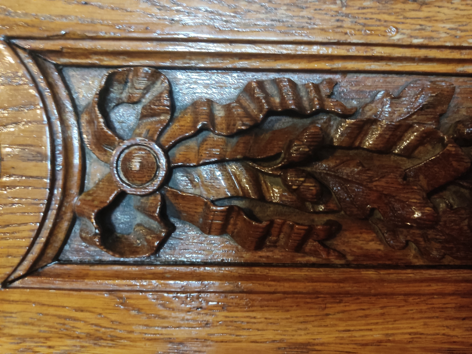 A close-up shot shows a section of detailed wood carving. The carving is set within a rectangular frame of wood and features a design including a bow with a circular centerpiece, followed by a trailing ribbon and oak leaves. The wood is a medium brown tone, and the carving has been finished with a glossy sealant. The underlying wood grain is visible. The image lacks contextual information about location or activity in Saint Petersburg.