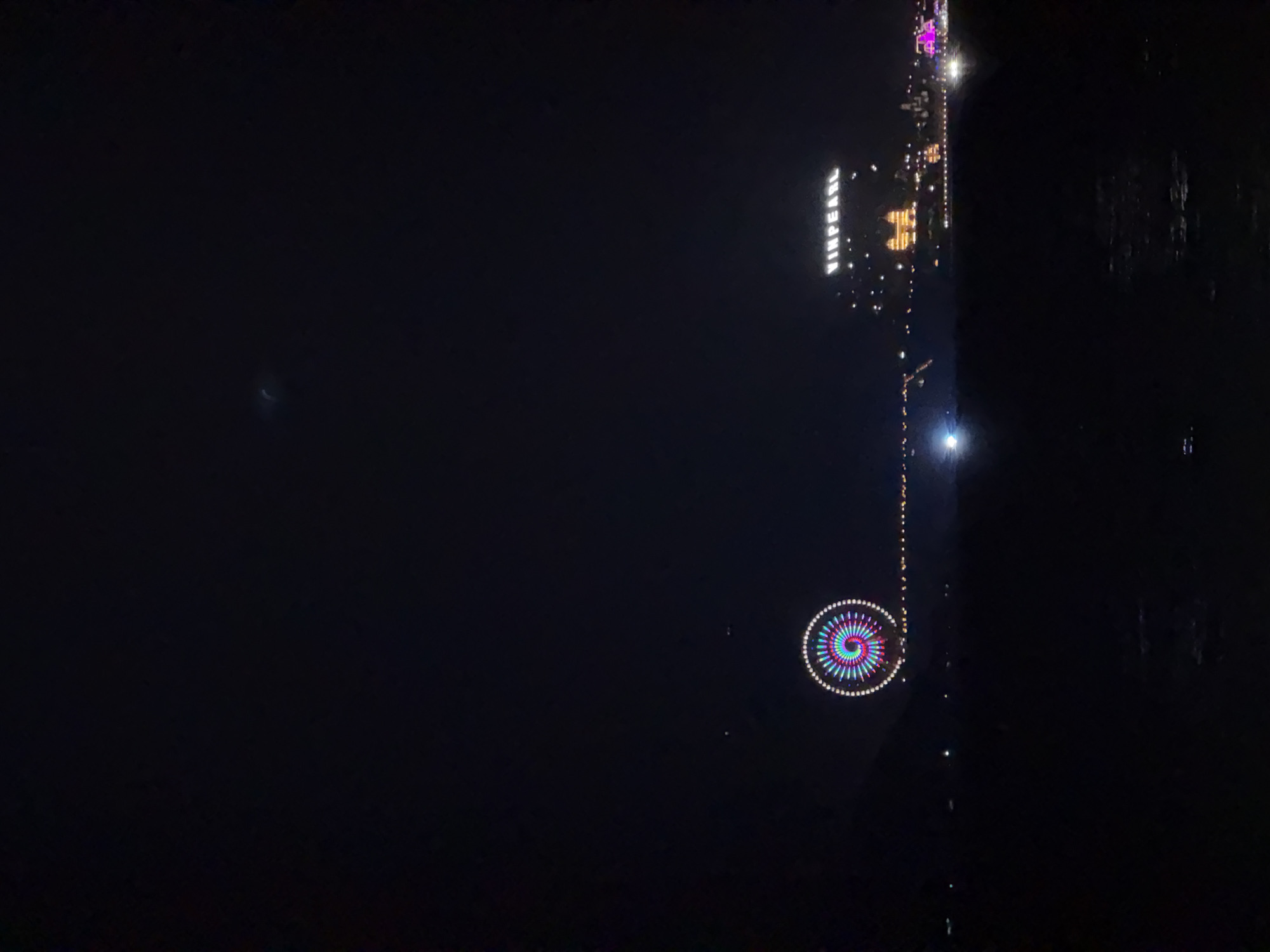 At night, the city of Nha Trang, Vietnam, is reflected in dark water. “VINPEARL” is spelled out in illuminated letters on a building on the right. Below, a string of lights leads to a Ferris wheel, lit with concentric rings of red, blue, purple, green, and white.