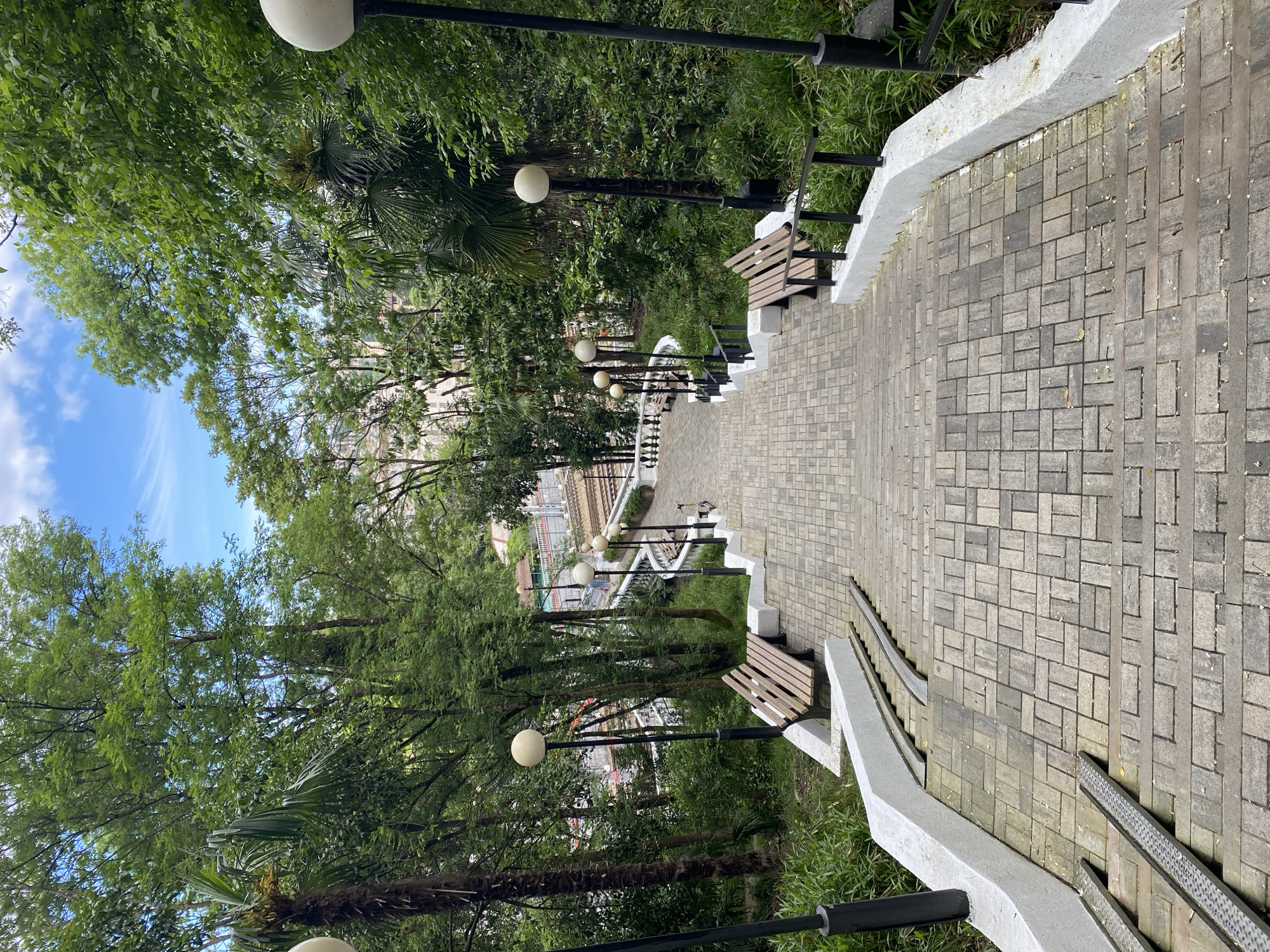The image shows a descending brick staircase in what appears to be a park or garden setting in Sochi, Russia. The staircase is bordered by white concrete walls on either side, with black metal railings in some sections. Benches are placed along the staircase. Lamp posts with spherical lights are positioned along the path, amidst trees and greenery. In the distance, buildings are visible between the trees. The sky is partially visible through the trees.