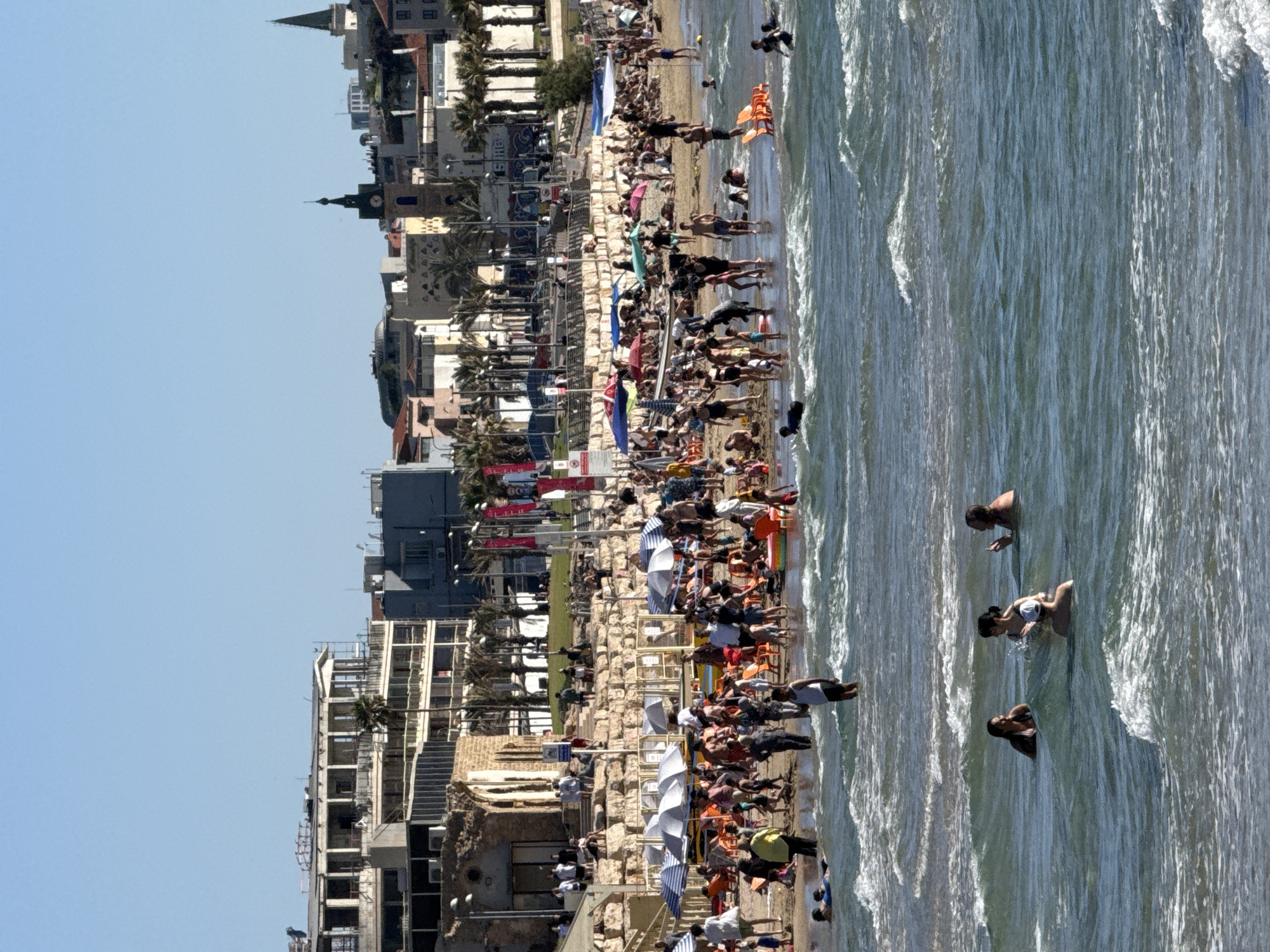 An overhead shot captures a vibrant beach scene in Tel Aviv, Israel, stretching from a bustling coastline to the calm sea under a clear blue sky. The image is oriented vertically, showing the horizon running down the center.
On the left, a dense urban landscape features a mix of modern and older buildings, characteristic of the Jaffa (Yafo) area of Tel Aviv. Several structures have distinctive architectural elements like spires and domed roofs, with many multistory buildings lining the coast. Palm trees are visible throughout the built-up area, adding to the Mediterranean atmosphere.
The central part of the image is dedicated to the sandy beach, which is densely populated with people. Many individuals are relaxing on lounge chairs, some under striped or colorful beach umbrellas, suggesting a sunny day. Others are standing or walking along the shoreline. Various beach amenities like kiosks or changing rooms are also visible.
To the right, the turquoise-green sea is filled with people swimming, wading, and enjoying the water. Gentle waves break near the shore, indicating calm conditions. The bright sunlight creates reflections and glints on the water's surface.
The overall impression is one of leisure and enjoyment on a busy beach day. The time of day appears to be mid-morning or early afternoon, given the bright light and clear shadows. No specific readable text is clearly discernible on signs or banners within the image.
