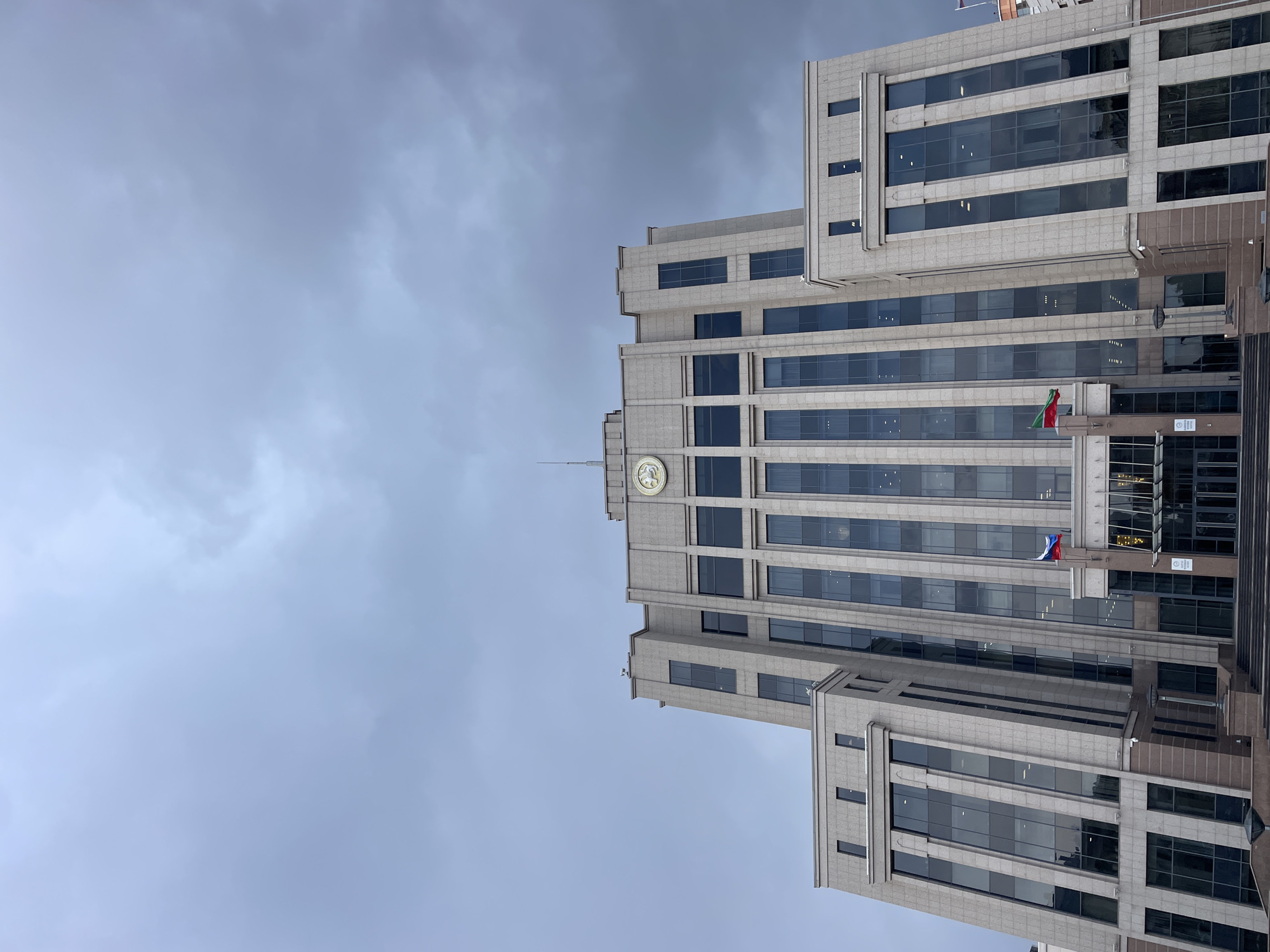 The image captures a tall, modern building in a portrait orientation, viewed from below against an overcast sky. The building, presumably located in Kazan, Russia, is constructed from light-grey stone or concrete and features numerous dark, reflective windows arranged in vertical strips and horizontal bands. Its architecture includes several tiered setbacks and projecting sections, creating a structured and imposing facade.
Near the top of the central section of the building, a golden circular emblem is visible, depicting what appears to be the double-headed eagle of the Russian Federation's coat of arms. Lower down on the building's facade, two flags are displayed. The upper flag shows horizontal green, white, and red stripes, representing the flag of Tatarstan. Below it, the Russian Federation flag, with horizontal white, blue, and red stripes, is flown. These flags serve as strong location cues, confirming the setting in Kazan.
The sky is uniformly cloudy and grey, suggesting an overcast day with diffused lighting, precluding any harsh shadows. There are no people visible, and no specific activity or event is taking place, presenting a static architectural study. No clearly legible text is discernible on the building's facade or signs.