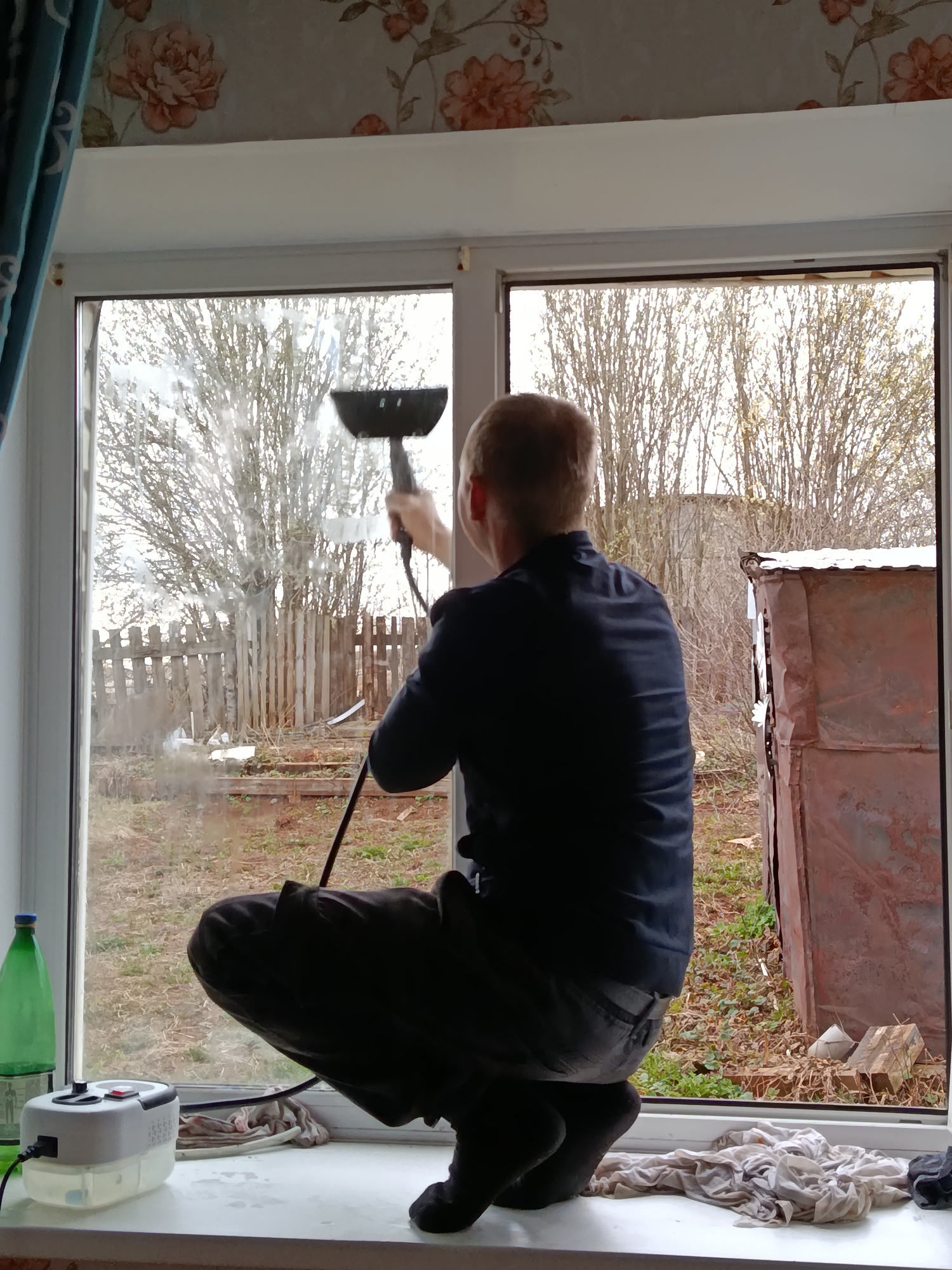 A person is shown cleaning a window from inside a building. The person is in a squatting position on a window sill, facing the window and holding a handheld cleaning device with a black handle and a wider black head. The window is partly cleaned, with streaks of moisture visible. Outside the window, the scene includes bare trees, a wooden fence, a metal structure, and ground covered with grass and leaves. On the window sill inside, there is a white and clear cleaning machine with a power cord, a green glass bottle, and folded pieces of fabric. The window frame is white, and the wallpaper above the window has a floral pattern. A blue curtain hangs to the left of the window.