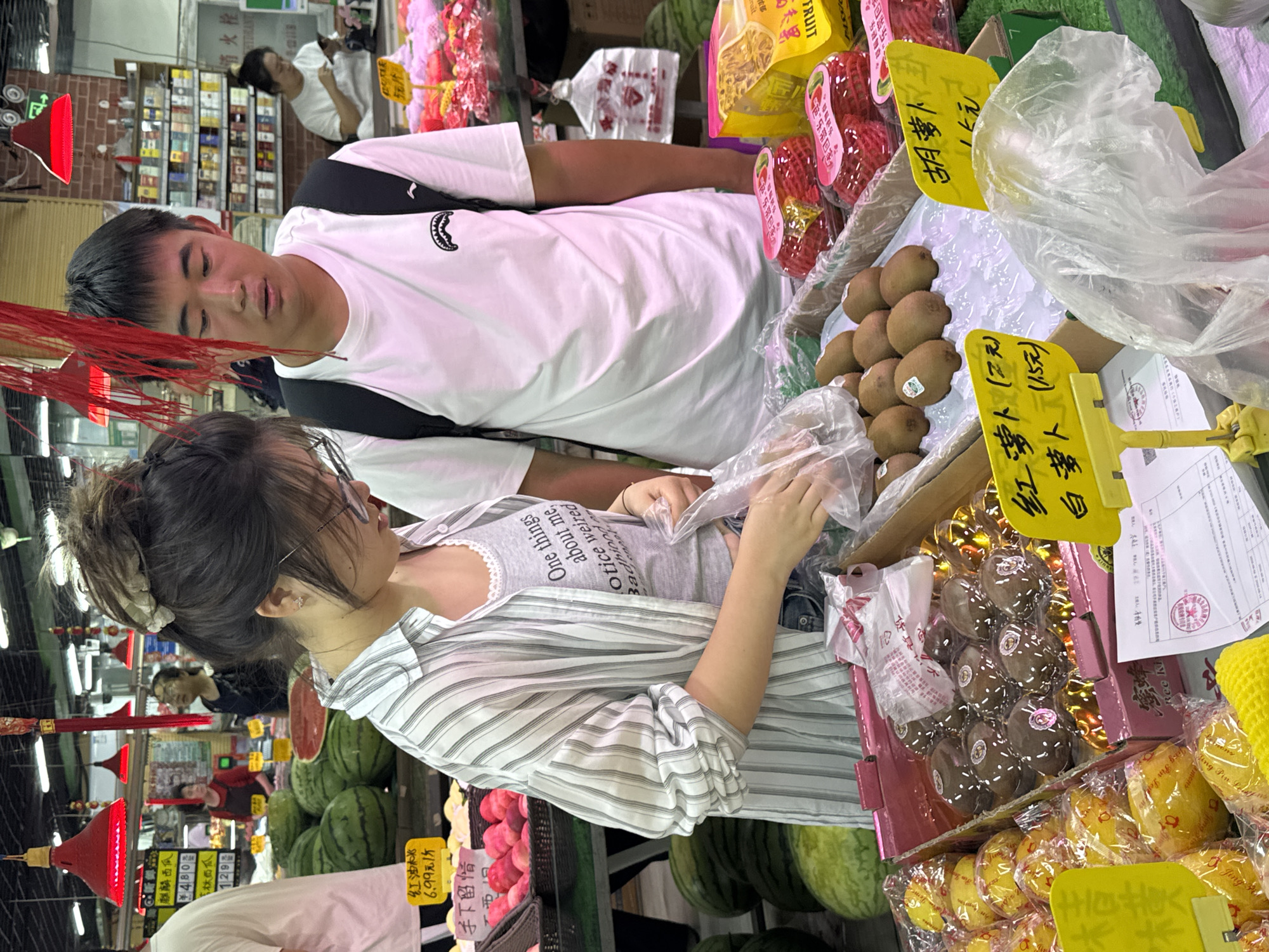 In the city of Zhongtun, China, a young couple is shopping for fruit at a market. The female, wearing glasses and a striped shirt over a gray tank top, is examining kiwis in a plastic bag. She has dark hair pulled up in a bun. The male, wearing a white t-shirt and a backpack, stands beside her. The market stall features various fruits, including watermelons, apples, and packaged kiwis, with price tags in Chinese. The background shows shelves stocked with goods and other shoppers.