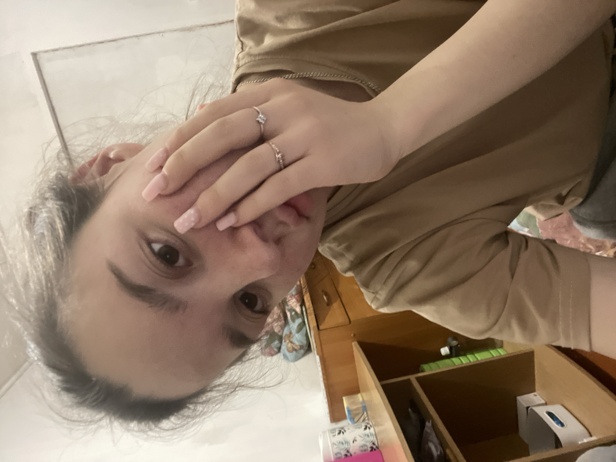 A woman is holding her hand to her face, partially covering it. She is wearing two rings. The background includes a shelf with various items, a bed or couch with patterned cushions, and a wall. There is no information available to determine the location of the image in Ольховка, Russia.