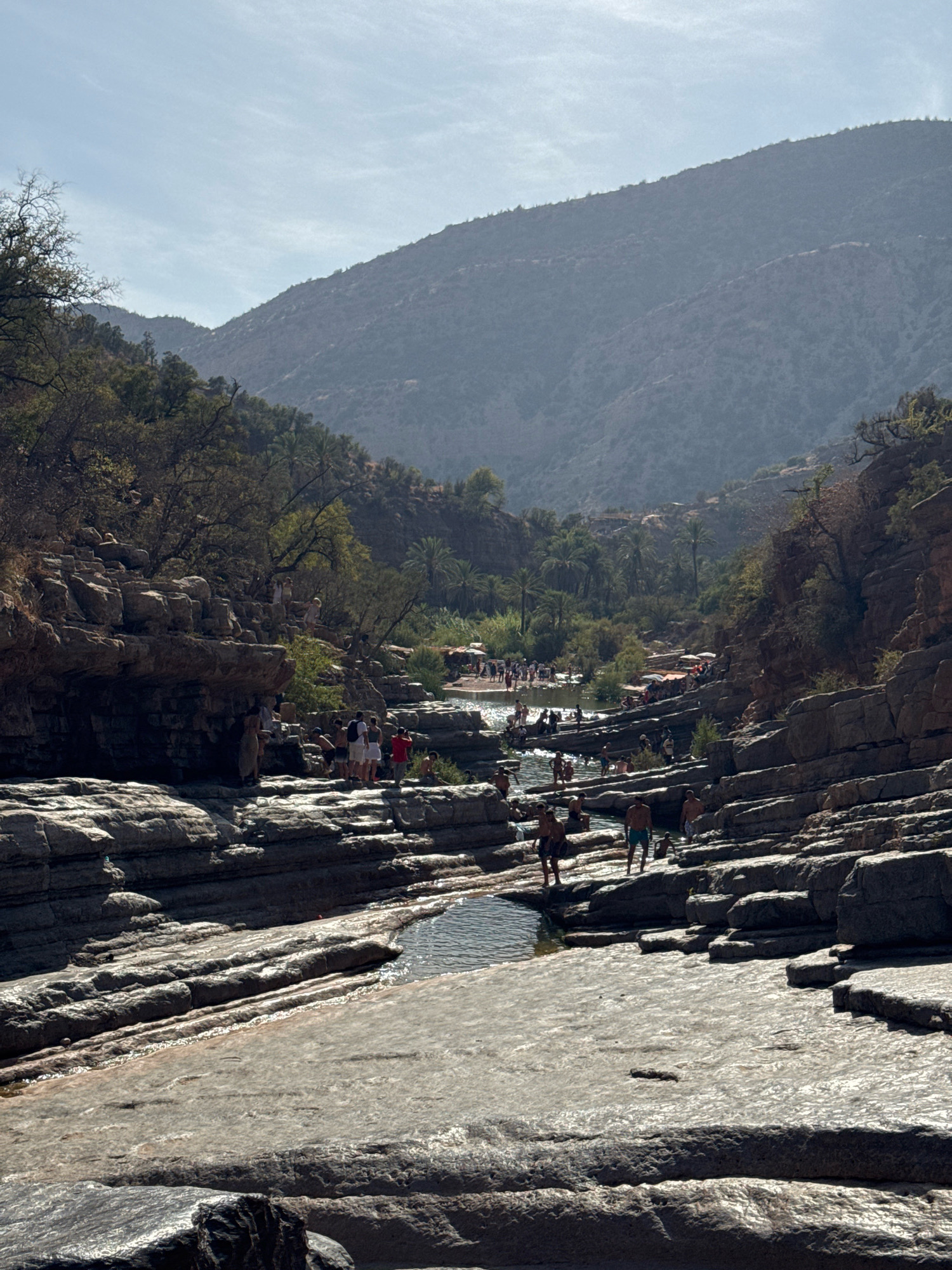 The image depicts a natural landscape with a river flowing through a rocky gorge. The terrain is characterized by layered rock formations and sparse vegetation, including trees and palm trees. In the distance, hazy mountains rise against a bright sky.
A significant number of people are present, scattered along the riverbanks and within the shallow water. They are engaged in various activities, including wading, swimming, and sitting on the rocks. The individuals are dressed in casual summer clothing, suggesting a recreational outing.
The scene appears to be a popular natural attraction, possibly a park or a designated swimming area. The presence of umbrellas and what seem to be seating areas on the right side of the image further supports this interpretation. The overall atmosphere is one of leisure and outdoor activity in a scenic, natural setting.
The location is identified as Aqesri, Morocco, a place known for its natural beauty and outdoor recreational opportunities. The image captures a vibrant day with many people enjoying the river and the surrounding environment.