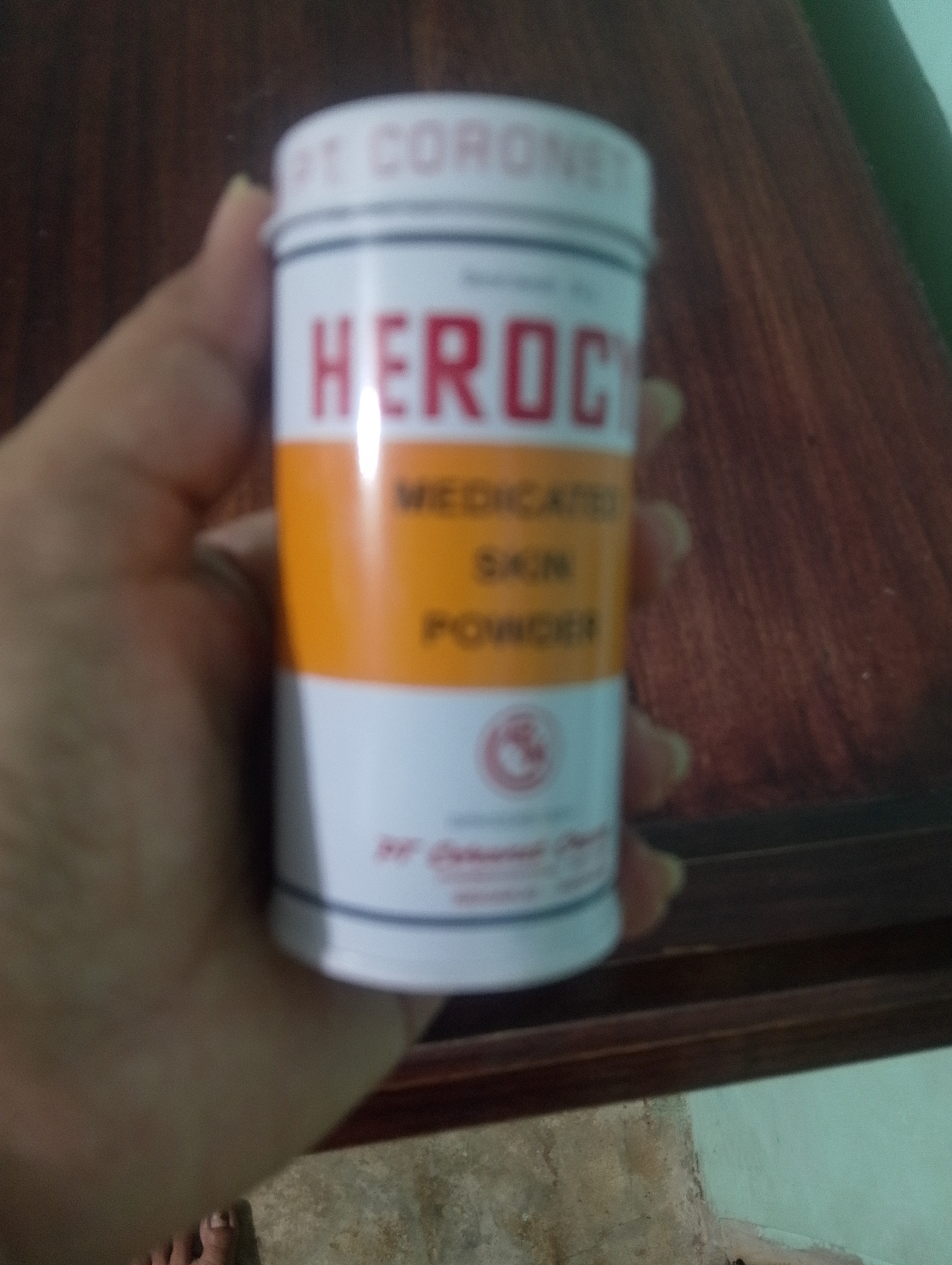 The image shows a hand holding a white and orange cylindrical container of "HEROCYN MEDICATED SKIN POWDER". The container has red lettering and a blue band. The background is a dark wooden surface with a greenish-grayish wall in the upper right corner and a textured gray floor in the lower portion. The image is blurry and taken from a slightly upward angle, obscuring details of the surroundings. The lighting is dim, casting a soft glow on the product. The setting is likely indoors, possibly a home or shop, in Serbelawan, Indonesia.