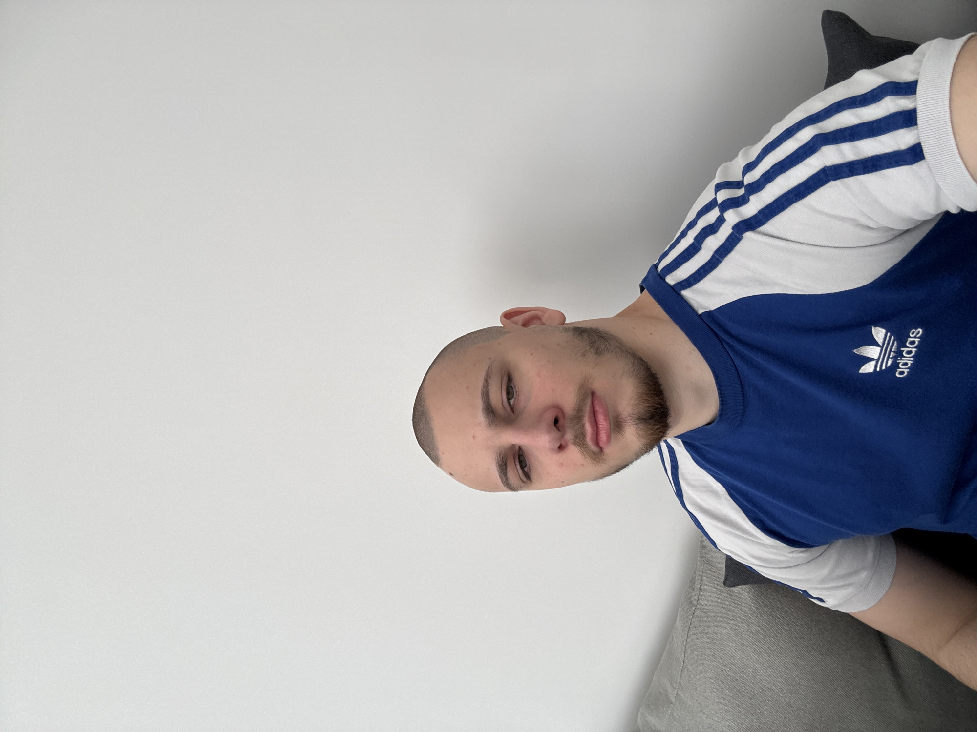 A fair-skinned man with a closely shaved head and a beard is wearing a blue Adidas t-shirt with white stripes on the sleeves. He appears to be lying down on a grey sofa. The background is a plain white wall. The location is not discernible in the image.