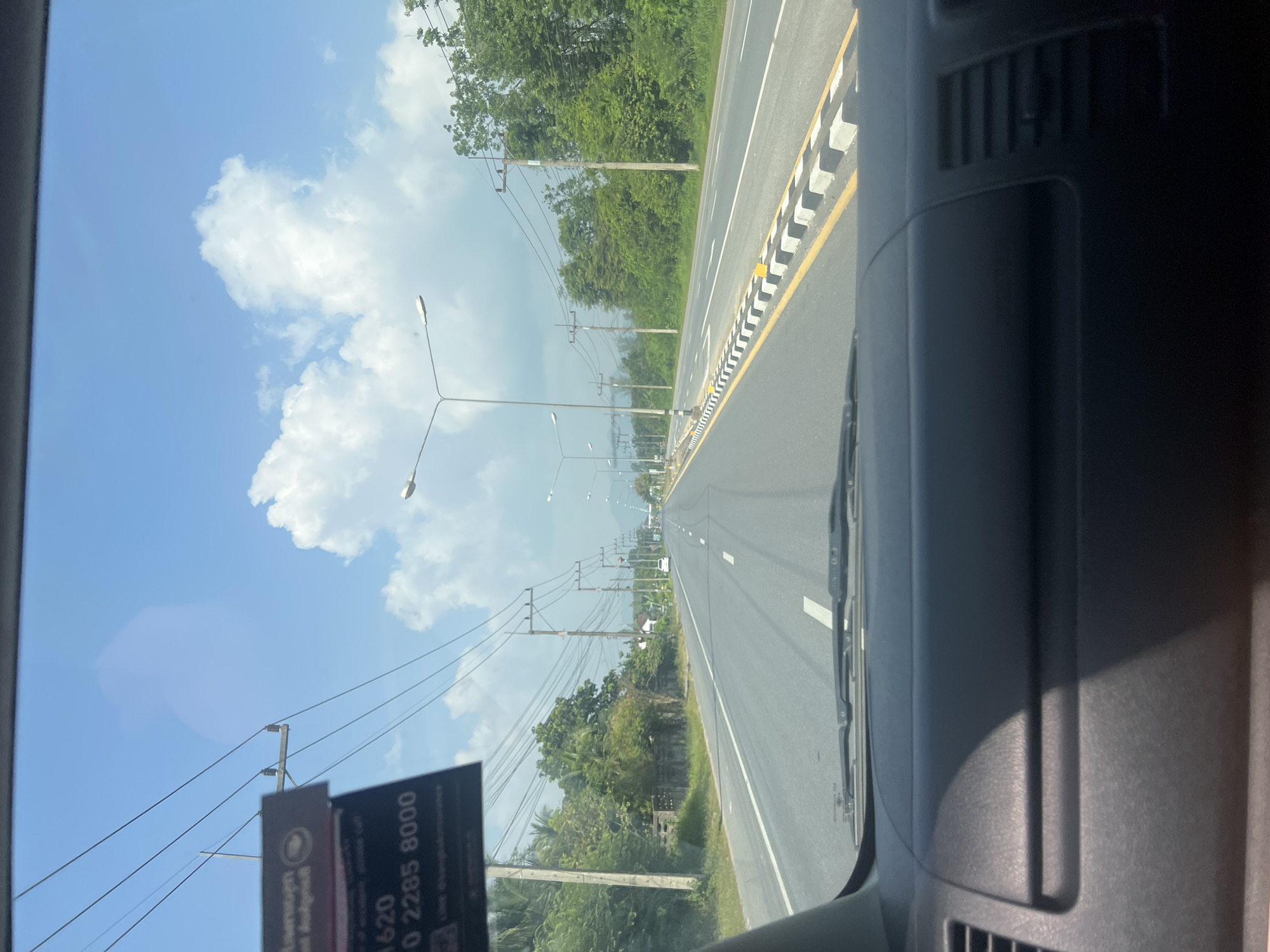 The image shows a view from the inside of a vehicle, looking forward through the windshield at a road in Khao Phra, Thailand. The road is paved and has multiple lanes, with white lane markings. On the right side of the road, there is a raised median with black and white striped markings. Trees and utility poles line the sides of the road. The sky is blue with scattered white clouds. In the distance, mountains are visible. The dashboard of the vehicle is visible in the foreground. A sticker is affixed to the windshield on the left side, with some text and numbers that are partially obscured.