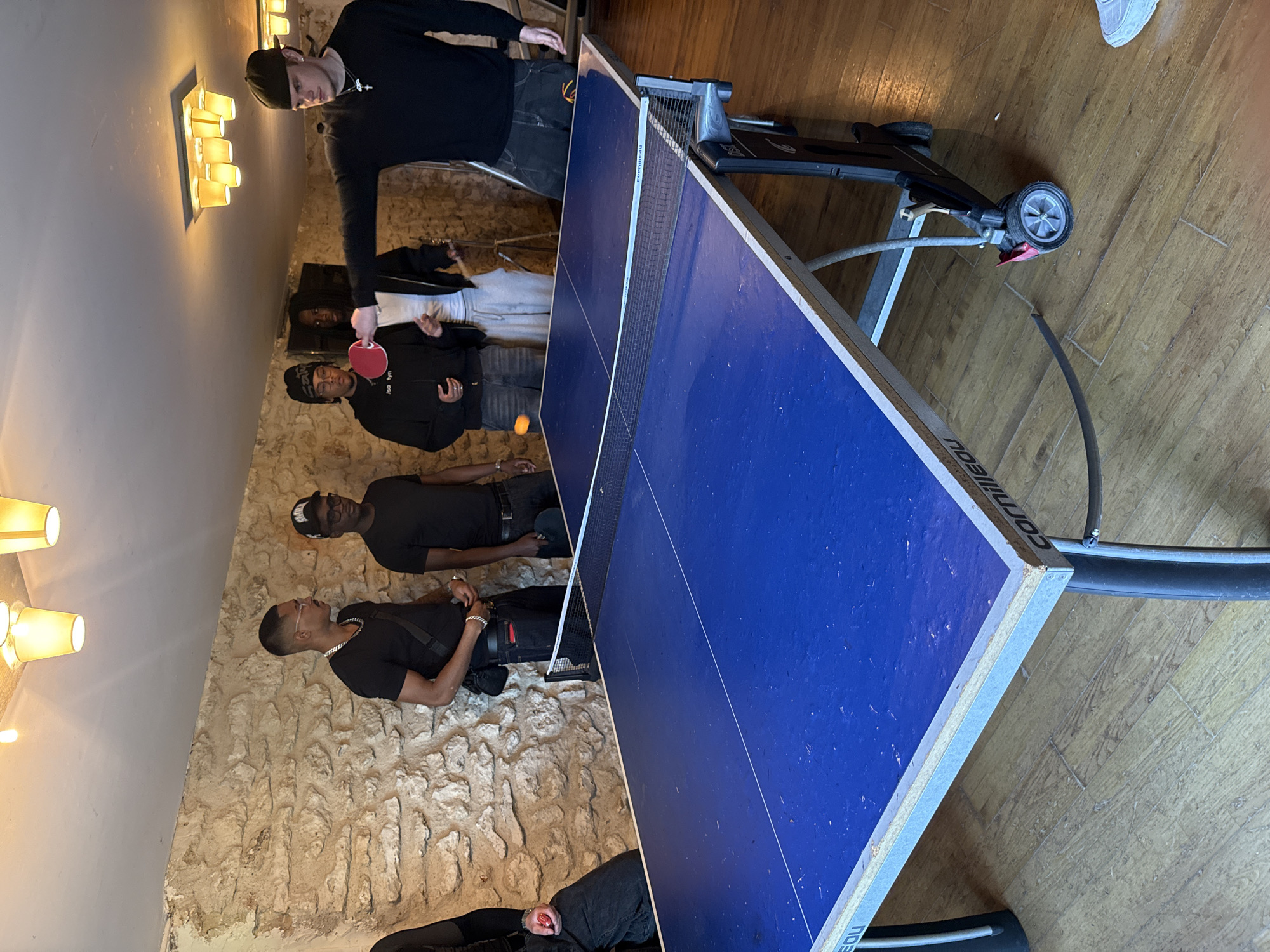 The image depicts a group of six people playing ping pong indoors. They are in a room with a textured wall, possibly stone, and wooden floors. The ping pong table is blue and has a net. One person is hitting the ping pong ball with a paddle, while others are watching. The image was taken in the city of Palaiseau, France. A scooter is parked near the table.