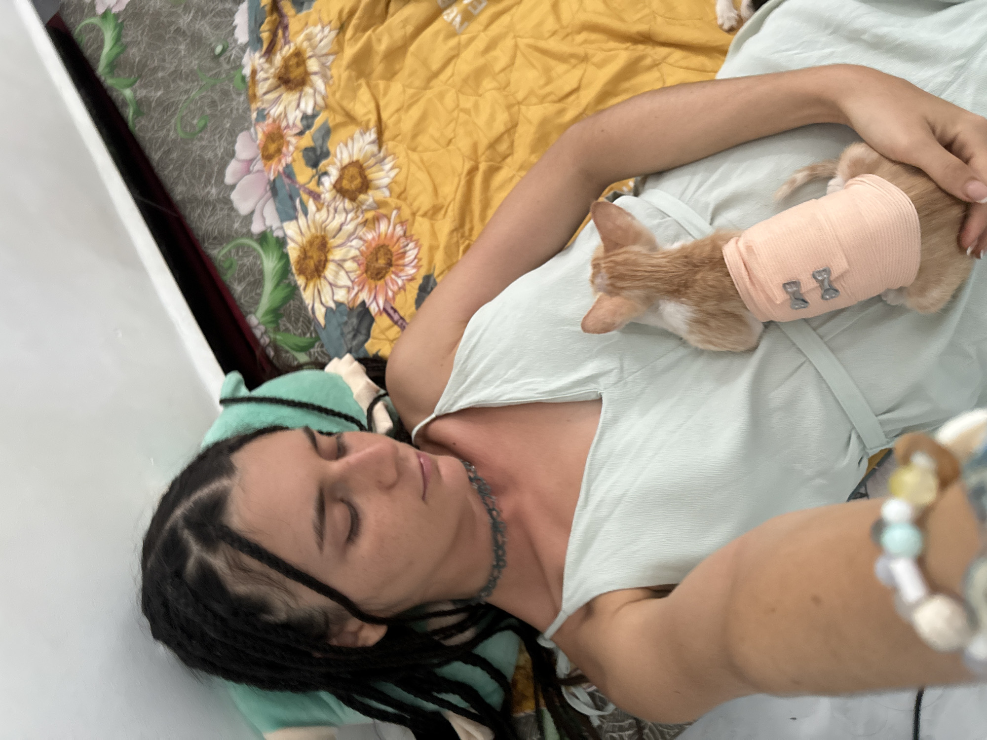 A young woman with dark dreadlocks lies on her back on a bed in a room in Mũi Né, Vietnam. She is wearing a light green slip dress and a beaded choker necklace. Her eyes are closed, and she is holding a small, orange and white kitten wrapped in a light pink bandage. The kitten is facing away from the camera, resting on the woman's chest. The bed is covered with a yellow blanket featuring a sunflower pattern and a green, floral-patterned sheet. A turquoise pillow is visible behind the woman's head. The overall scene suggests a moment of rest and care for the injured kitten.