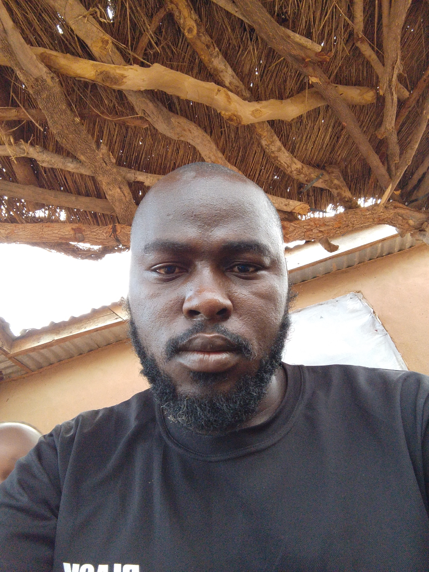 The image shows a close-up selfie of a Black man with a shaved head and a beard. He is wearing a black t-shirt. The background reveals a roof constructed of wooden branches and straw-like material. Beyond the roof, there is a glimpse of a building with corrugated metal. The location is Mulke Barikin Sayejo, Nigeria.
