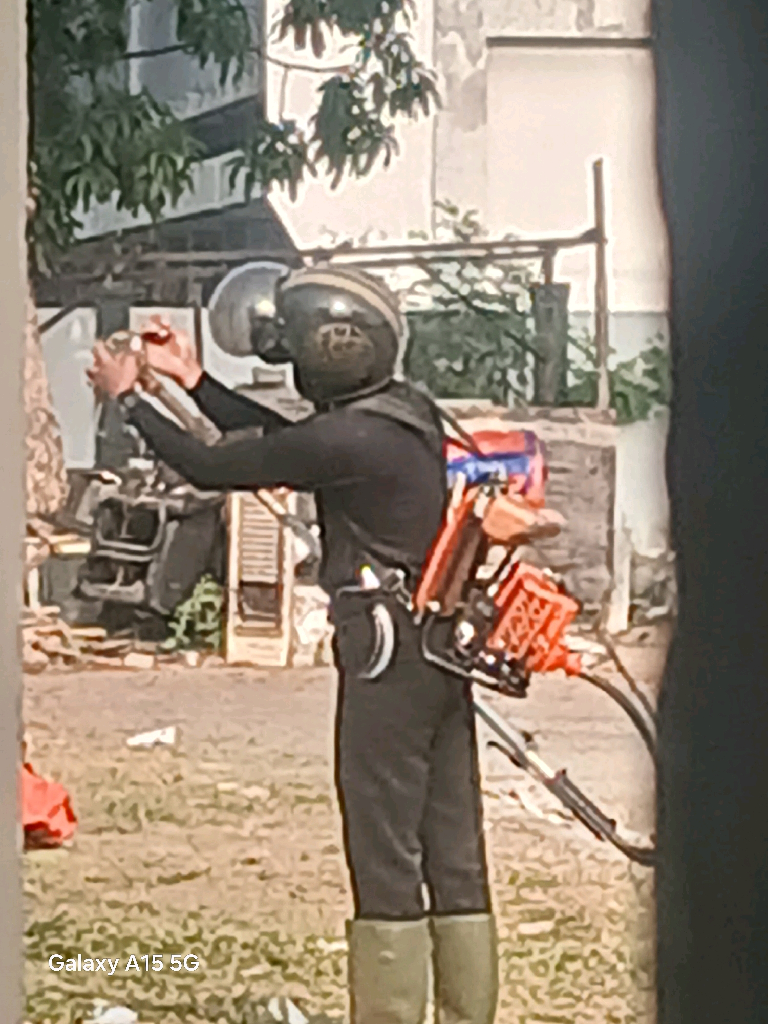 In Surabaya, Indonesia, a person wearing a black long-sleeved shirt, black pants, and green boots stands in a grassy outdoor area. They have a helmet on their head and are holding a device with a red and blue casing on their back. The person is also holding a metallic object in their hands, seemingly preparing to use it. In the background, there are buildings with visible windows and trees with green foliage. Various objects, including what appears to be machinery and debris, are scattered in the background. The overall setting suggests an urban or semi-urban environment with signs of disarray or construction.