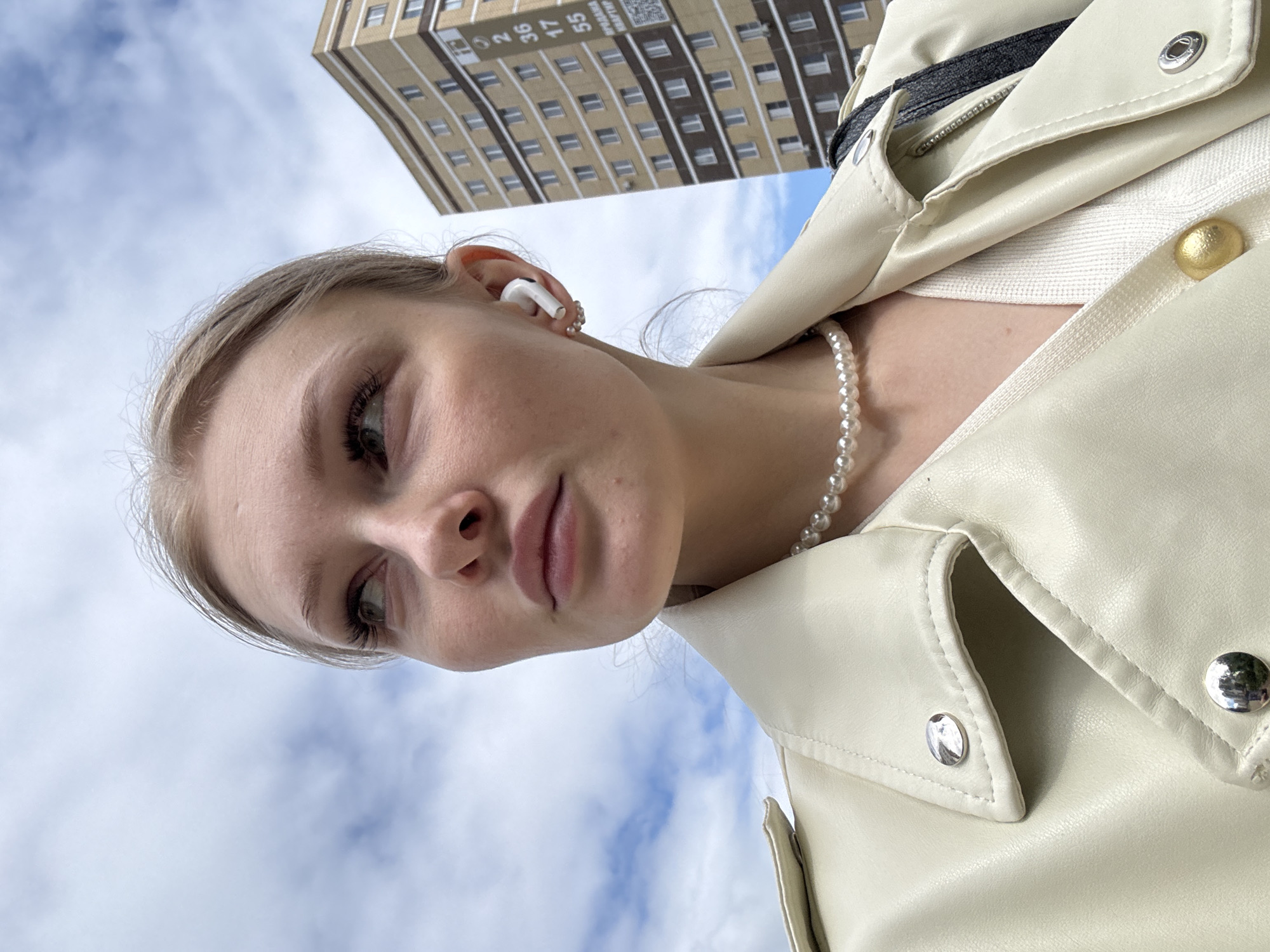 A young woman with light blonde hair pulled back is looking upwards at the camera. She is wearing a white wireless earbud in her left ear and a pearl necklace. She is also wearing a light-colored leather jacket. Behind her and to the left is a tall building with a repeating pattern of windows, indicative of an apartment complex. The building's facade is a light beige or tan color with dark brown accents around the windows. There is text visible on the building that says "ПЕРМЬ" and "KBAРТИРА" in Russian, indicating this scene is in Perm, Russia. Above and to the left of the woman is a blue sky with scattered white clouds. The photo appears to have been taken from a low angle, looking up at the subject and the building.