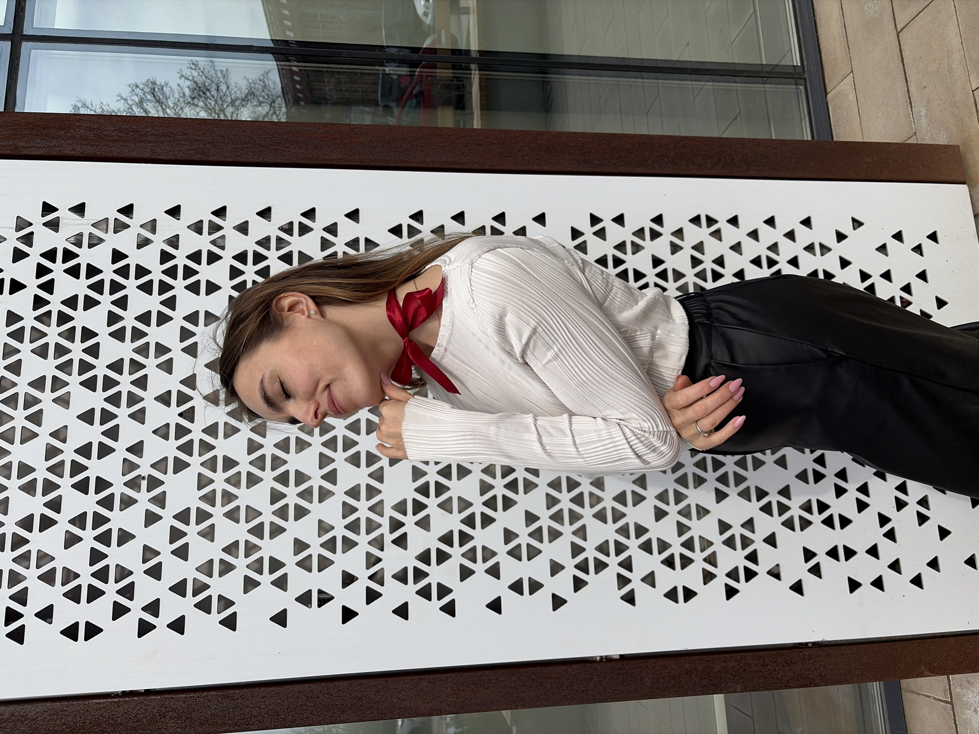 A young woman is lying on a white metal bench in an urban setting. She is wearing a white long-sleeved shirt, black pants, and a red ribbon tied around her neck. Her head is resting on the bench, and her eyes are closed. The bench has a pattern of triangular cutouts. A window with a reflection is visible in the background. The location is Izhevsk, Russia.