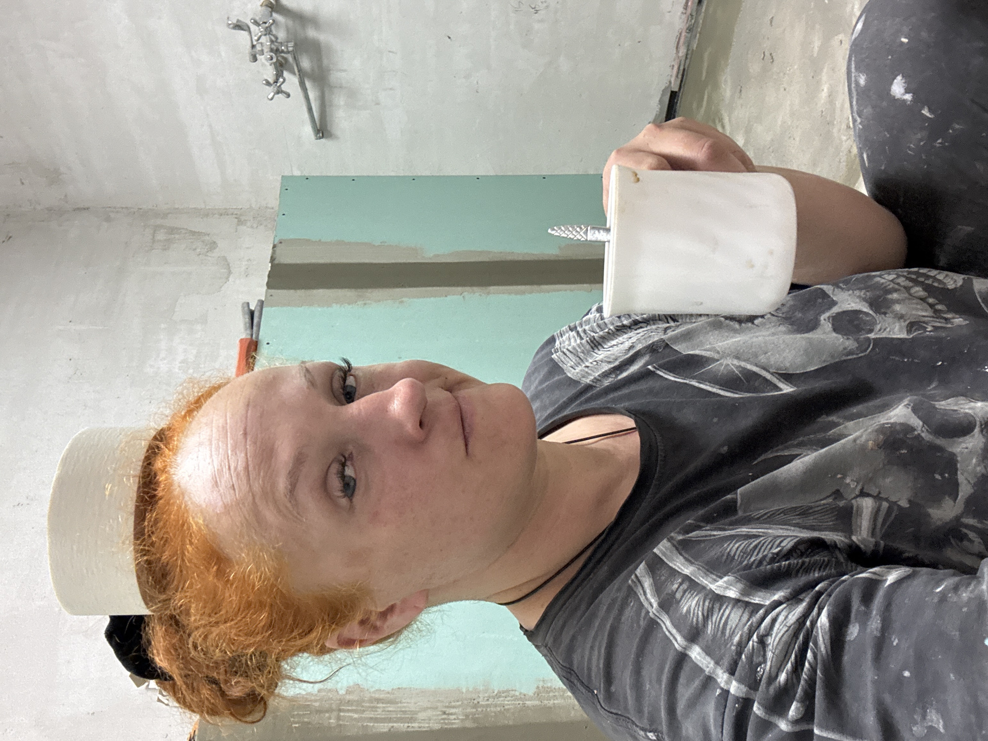 A woman with red hair poses indoors. She is wearing a black t-shirt and holds a white object with a metal drill bit sticking out of it. A roll of white material sits on her head. The room behind her shows unfinished drywall and plumbing fixtures. The location is Chaikovsky, Russia.