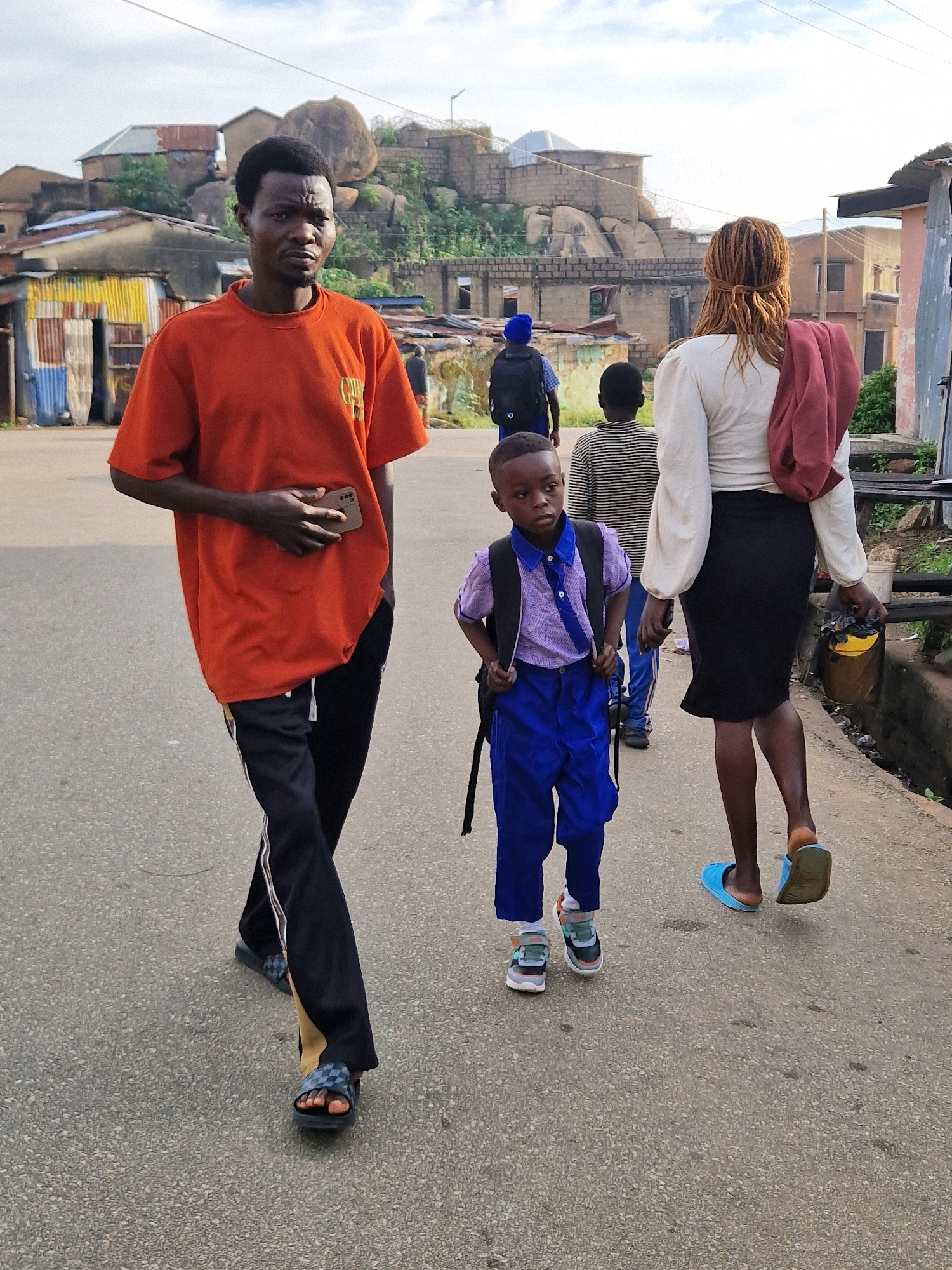 In the city of Jos, Nigeria, a group of people walk along a dusty, paved road. In the foreground, a man in an orange t-shirt and black track pants walks towards the viewer, holding a smartphone. To his right, a young boy in a blue uniform and backpack walks ahead of him, looking to the side. Behind the boy, a woman in a white shirt and black skirt with dreadlocks tied back, carries a red cloth over her shoulder and a yellow bucket in her hand. Further in the background, another person in a blue shirt and backpack is visible. The environment is characterized by uneven, rocky terrain and buildings with corrugated iron roofs, suggesting a developing urban or semi-urban area. The sky is overcast.