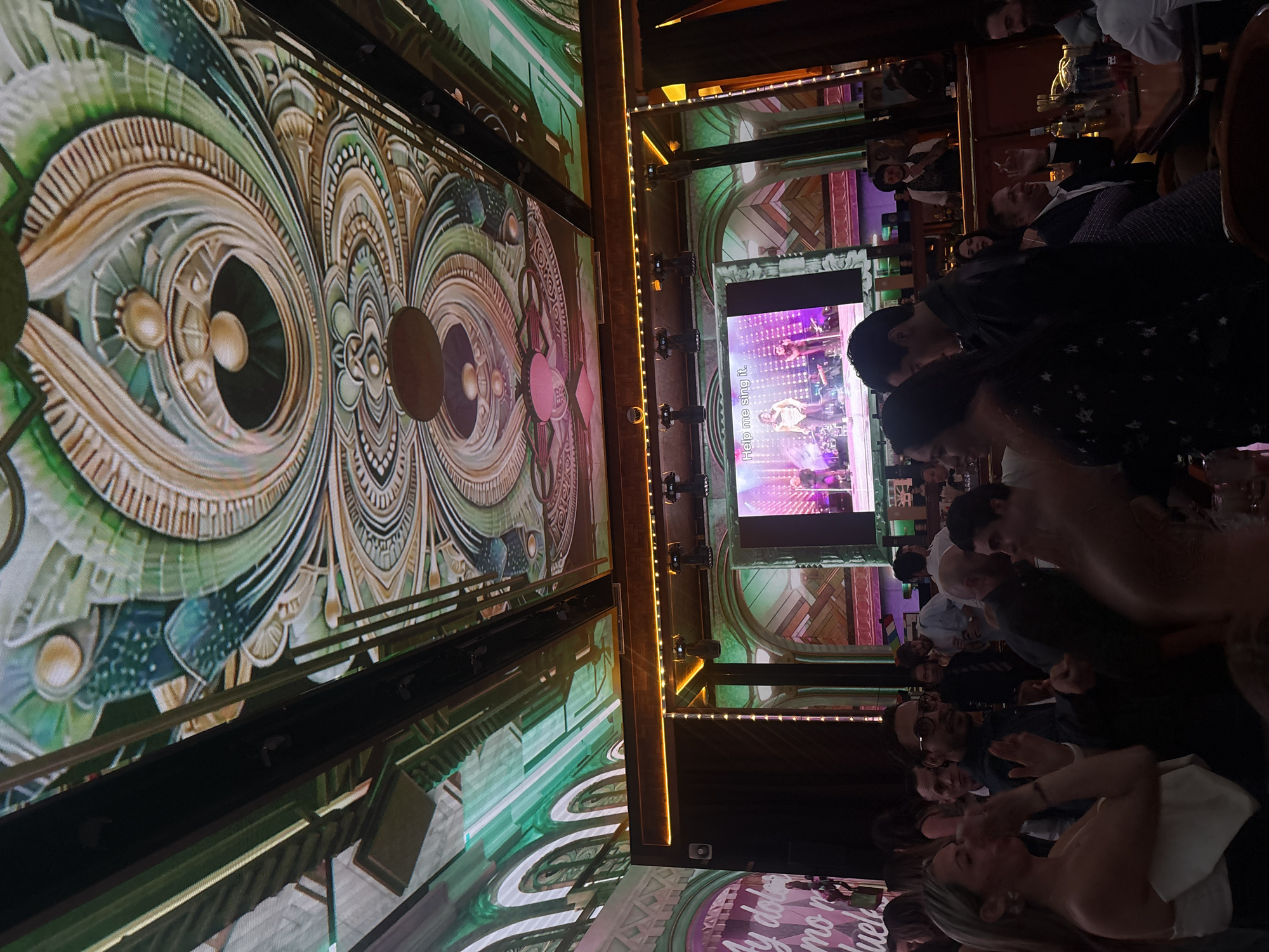 This image captures a vibrant nighttime scene inside an entertainment venue in Mexico City, Mexico, possibly a bar or a karaoke lounge. Numerous people are gathered, seated at tables, predominantly facing towards various screens and a central stage area, though the stage itself is not fully visible.
The setting is characterized by elaborate digital displays forming the walls. On the left, a large screen features an intricate, ornate pattern resembling Art Deco or Art Nouveau design, with swirling lines and geometric shapes in shades of green, gold, and white. On the right, a prominent screen displays what appears to be a musical performance or a karaoke video, brightly illuminated with purple and blue lights. English text, "Help me sing it.", is clearly visible on this screen. Another screen in the lower foreground on the left shows partially visible Spanish text, "Y duelen, no mue...", likely lyrics or a message.
The patrons are a mix of individuals and groups, appearing engaged and social. Some are seen clapping or looking intently at the screens, suggesting active participation in an event or performance. The lighting is primarily from the screens and decorative strip lights running along architectural beams, contributing to a lively and festive atmosphere typical of an evening out. The overall scene conveys a sense of entertainment and enjoyment among the attendees.