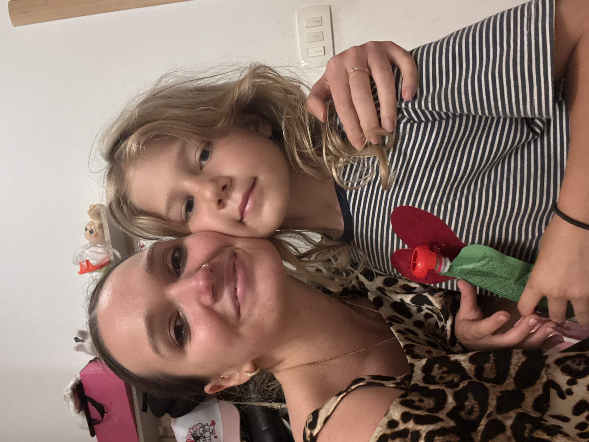 In an indoor setting, possibly a room in Budva, Montenegro, a woman and a child are captured in a close-up selfie. The woman, on the left, has her arm around the child and is leaning her head towards the child's cheek. She is wearing a leopard print top and a gold necklace. The child, on the right, has blonde hair and is wearing a striped t-shirt with a red heart applique. The child is holding a green pouch with a red cap in their hands. The background includes shelves with various items, including a pink box and figurines. A light switch with three buttons is visible on the right side of the frame. The overall scene depicts a moment of affection between the two individuals.