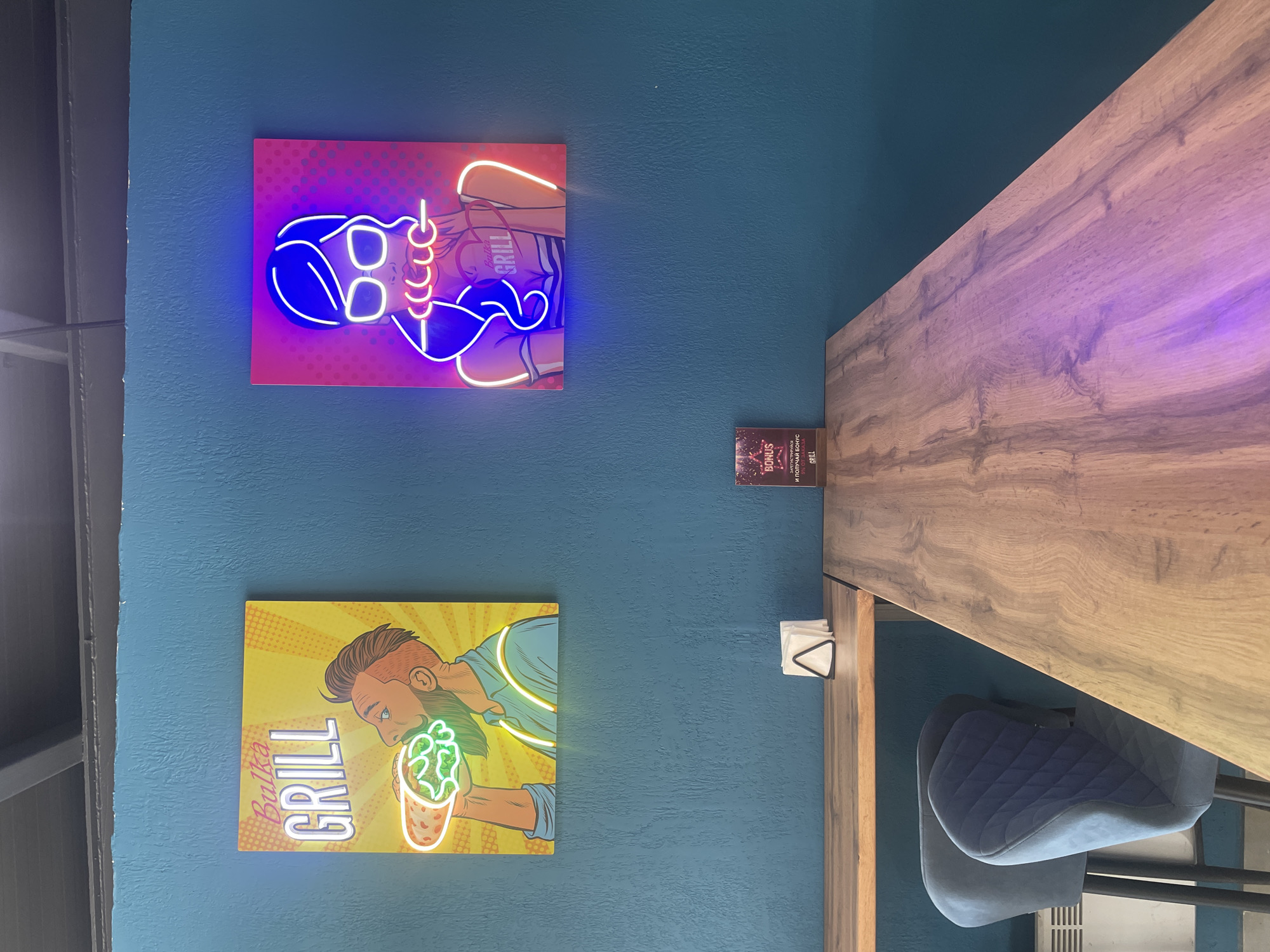 The image shows a portion of an interior, likely a restaurant or eatery in Krasnoyarsk, Russia. The wall is painted a teal color. On the wall are two framed neon-style artworks depicting cartoon figures with food themes. The upper artwork shows a woman with blue hair and glasses, wearing a top that reads "Grill". The lower artwork shows a man eating a food item with green fillings. To the right of the wall, there is a wooden countertop with a chair pulled up to it. A napkin holder sits on the countertop next to the wall. Between the artworks and the countertop there are bonus cards. The scene has a modern and casual aesthetic.