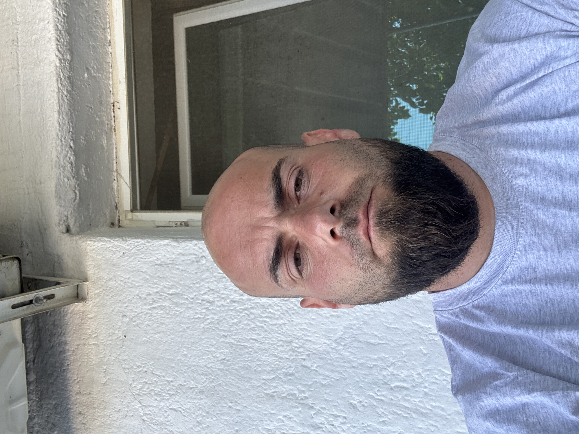 A head-and-shoulders portrait of a Caucasian man with a shaved head and full beard, taken outdoors. He is wearing a light gray t-shirt. The background includes a white textured wall, a window frame, and glimpses of foliage through the window. Specific location in Sarnevo, Bulgaria, cannot be determined from the image.