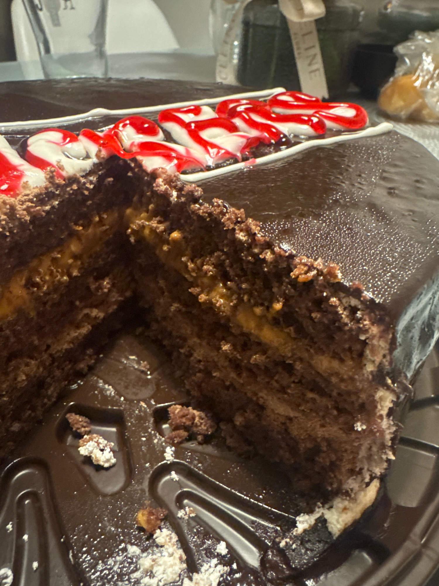 The image shows a close-up of a partially eaten chocolate cake with a glossy chocolate glaze and a filling that appears to be caramel or a similar sweet spread. The cake is decorated with red and white swirled icing that resembles candy canes. The cake is presented in a brown plastic container, and there are crumbs scattered within the container. The background is blurred, but it appears to be an indoor setting with some objects that are out of focus. There is no visible indication of the city of Almaty, Kazakhstan, or any specific location within it. The scene is a still life of a dessert.