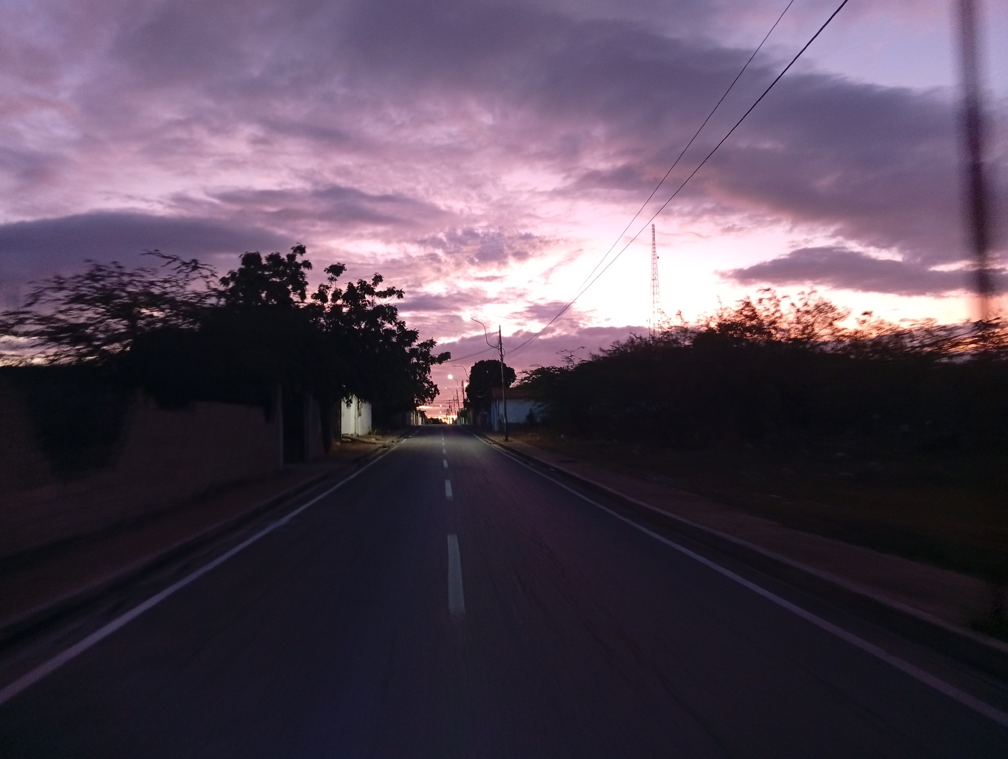 The image captures a twilight scene from a low-angle perspective, looking down a straight asphalt road in Juan Griego, Venezuela. The road features a white dashed center line and solid white lines along its edges, receding into the distance. The foreground and immediate sides of the road are dark, suggesting low light conditions.
Above, the sky is a dramatic canvas of clouds painted with vivid hues of purple, pink, and orange, indicative of either sunset or sunrise. The brightest light emanates from the horizon, illuminating the undersides of the clouds. Silhouetted trees and dense bushes line both sides of the road, appearing dark against the colorful sky. On the left, low, dark structures or walls are partially visible behind the vegetation. On the right, a tall, slender communication tower rises against the twilight sky in the mid-distance. Overhead, power lines stretch across the frame.
A faint light, possibly from a streetlight or distant building, can be seen in the far distance along the road. The scene evokes a sense of serene quietness. There are no people or vehicles visible, and no discernible text.