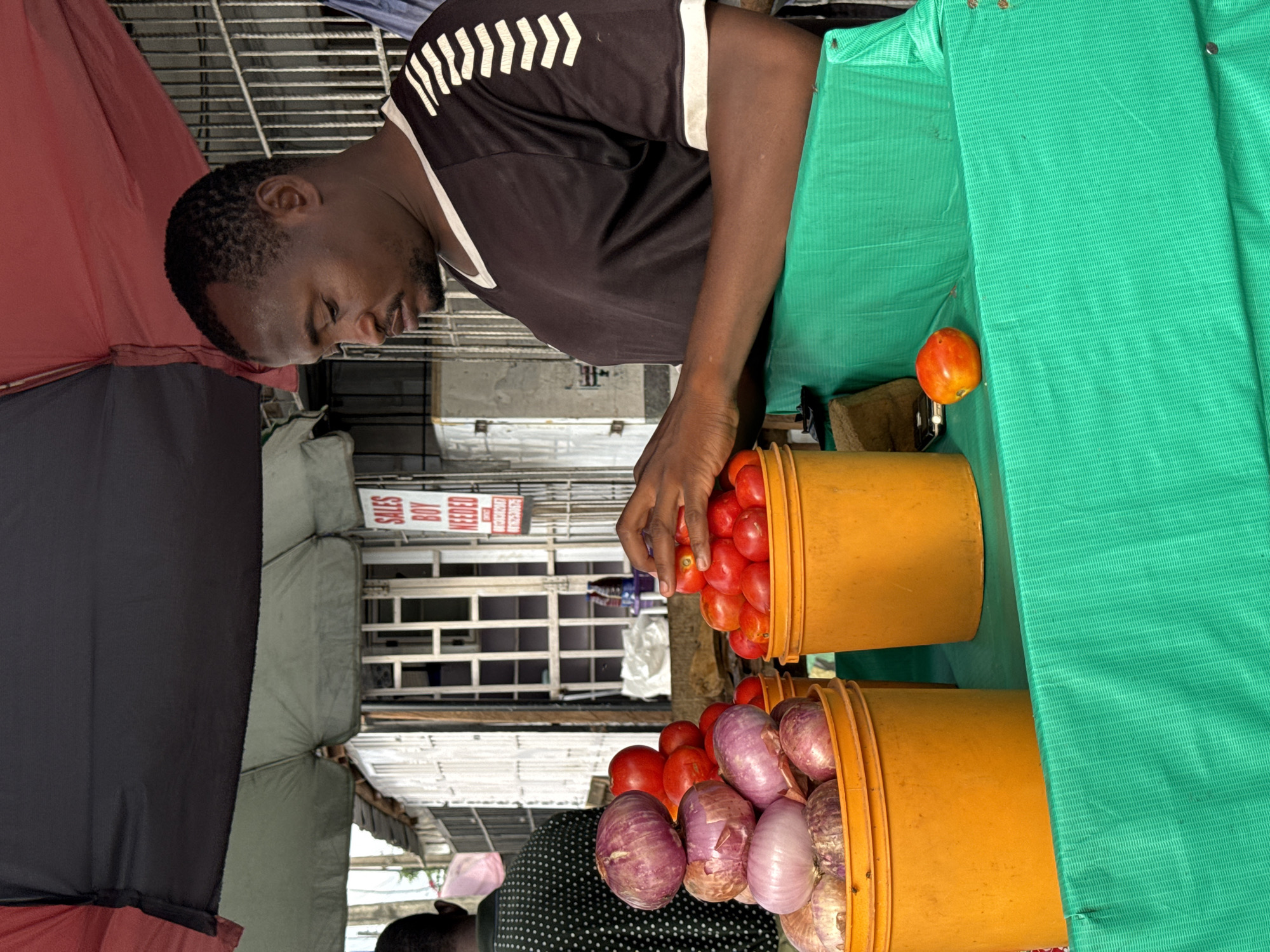 A street vendor in Ado, Nigeria, is shown in the image. The man is dark-skinned with short, dark hair and a trimmed beard. He is wearing a black athletic shirt with white stripes on the shoulder. He is behind a table covered in a green tarp. On the table, there are two orange buckets filled with produce. One bucket is full of tomatoes, and the man is picking through them. The second bucket is filled with red onions and some tomatoes. A single tomato sits on the green tarp. An umbrella provides shade for the vendor and his goods. In the background, there is a building with a sign that reads "SALES BOY NEEDED".
