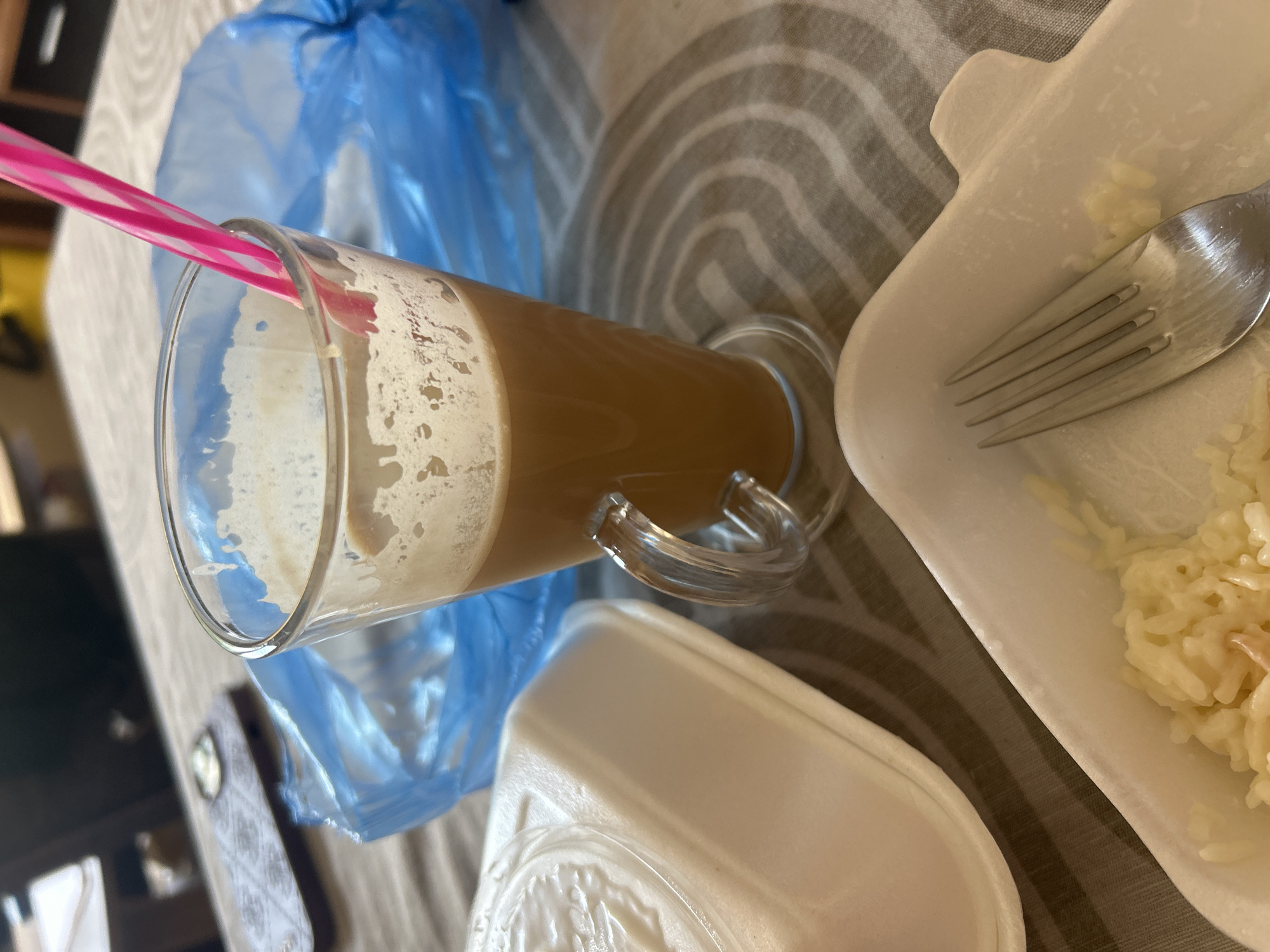 The image captures a close-up, high-angle view of a dining table setting, likely in Plovdiv, Bulgaria. The primary subjects are a tall glass filled with a light brown beverage, possibly coffee or a milkshake, topped with foam, and a pink and white striped straw. The glass has a curved handle. To the right of the drink, a white takeout container holds a portion of rice. A silver fork rests on the edge of this container. In the background, a bright blue plastic bag is partially visible, and the table is covered with a textured tablecloth featuring curved, grey stripes. The lighting suggests it is daytime, but no specific weather conditions can be inferred. The scene appears casual, possibly a meal taken at home or outdoors. There is no visible text in the image.