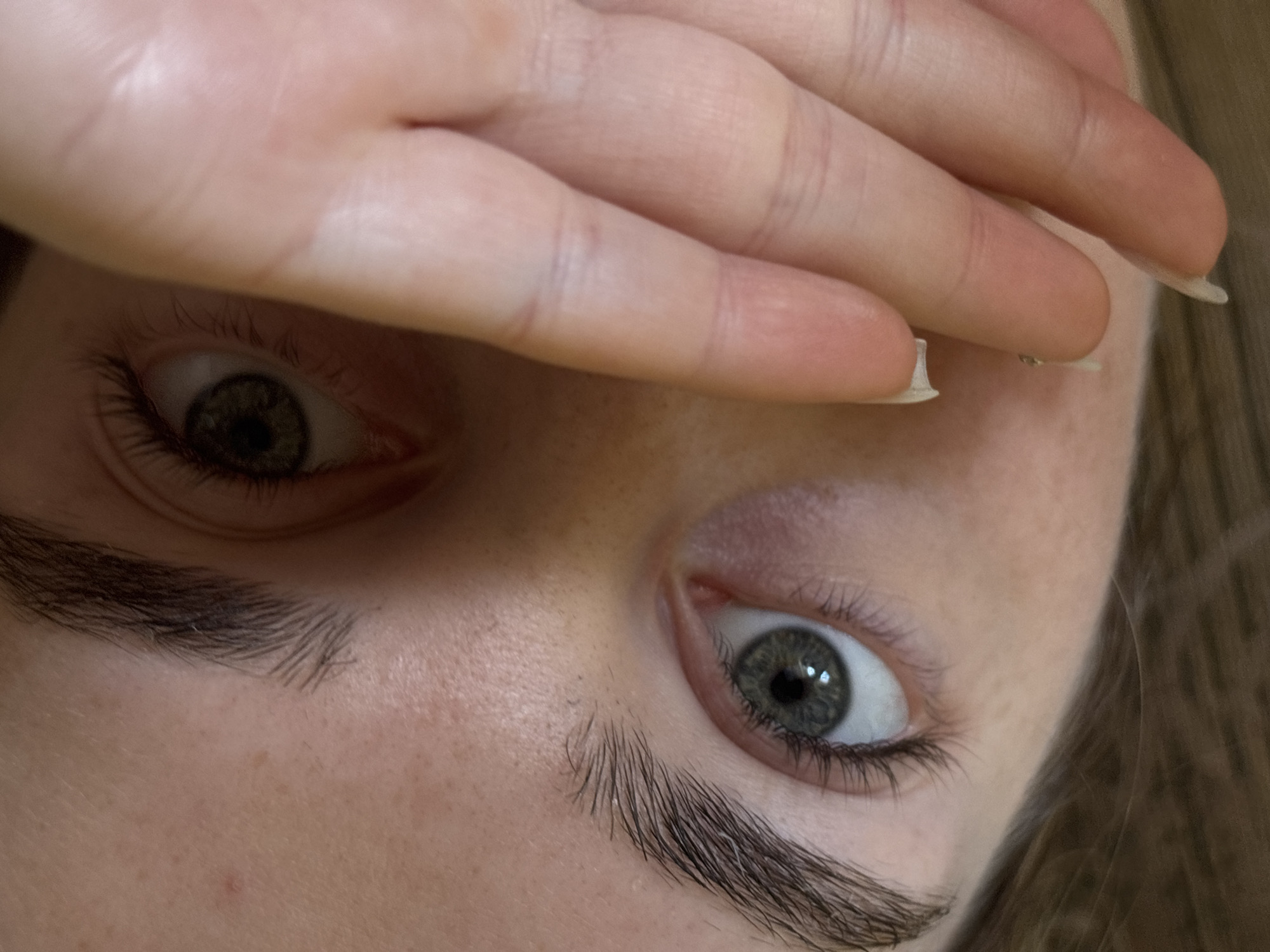 A close-up, upward-angled view captures the eyes and part of the face of an individual. A hand partially obscures the forehead, with light-toned skin visible on both the hand and face. The eyes are wide open, displaying green irises. Visible eyebrows are dark and relatively thick. A few manicured nails are also visible. The background is indistinct. No city is known or visible.