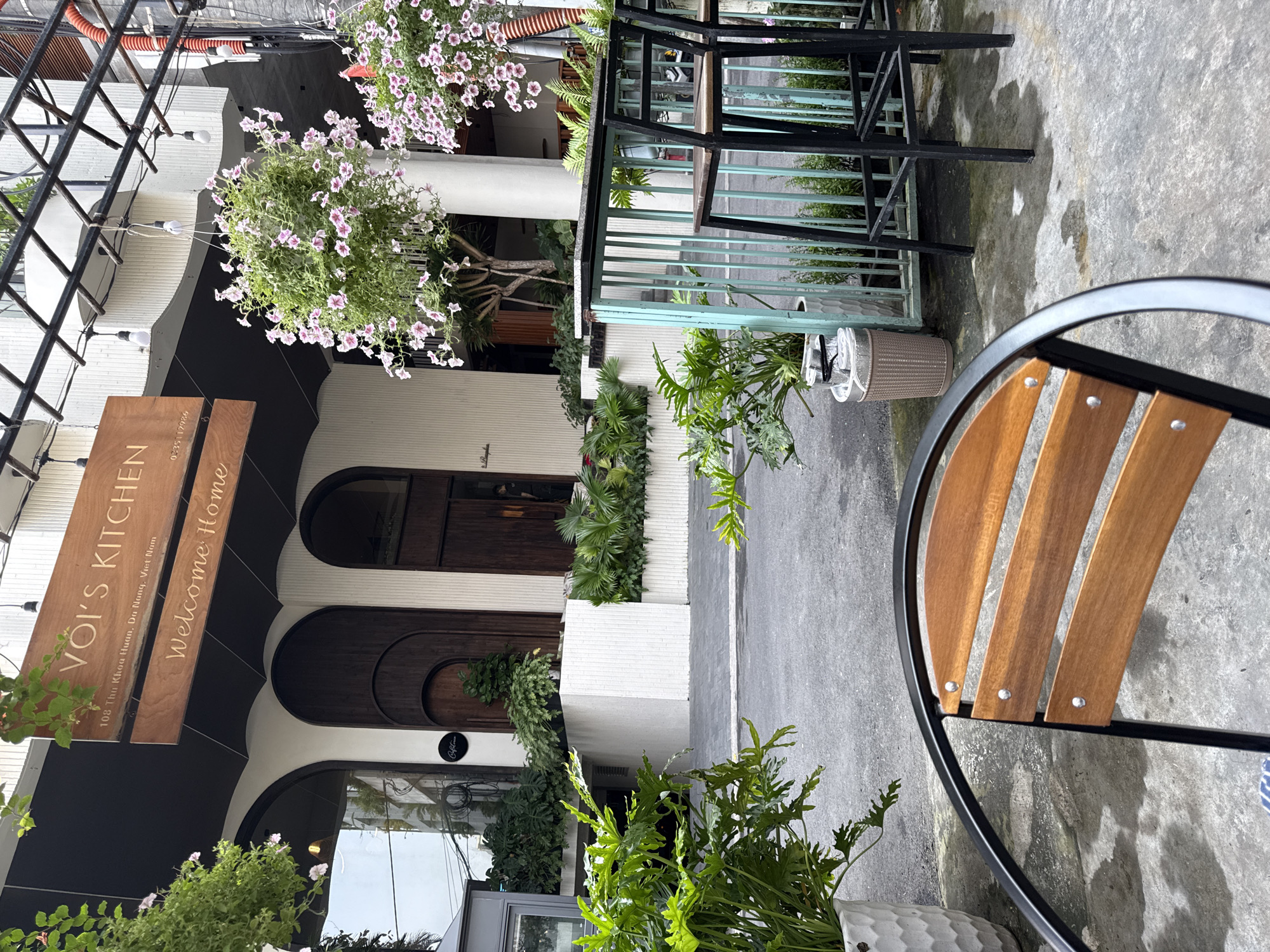 The image shows an outdoor setting with a focus on a cafe or restaurant called "VOI'S KITCHEN" located at 108 Thu Khoa Huan, Đà Nẵng, Vietnam. The building features a modern design with arched windows and doors, complemented by hanging plants with lilac flowers and greenery along the facade. A wooden sign displays the establishment's name and the phrase "Welcome Home." The foreground includes a wooden-backed metal chair. A metal railing separates the outdoor area from what appears to be a pedestrian walkway. The area appears to be wet, possibly from recent rain. There are no people visible.