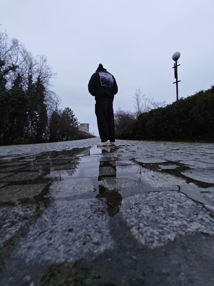 A low-angle image depicts a solitary individual standing on a wet, cobblestone pathway at the Monument to 1300 Years of Bulgaria in Shumen, Bulgaria. The person is viewed from behind, wearing a dark-colored hoodie featuring a rectangular, multi-toned grey and purple graphic on the back, dark pants, and light-colored sneakers. Their reflection, along with bare tree branches, is visible in a prominent puddle on the pathway in the foreground. To the left, dark, leafless trees are visible, partially obscuring a large, angular concrete structure characteristic of the monument complex. To the right, a dense, dark green hedge borders the pathway, behind which a tall pole topped with a spherical white light fixture is present. The sky is overcast, and the ground exhibits dampness from recent precipitation.