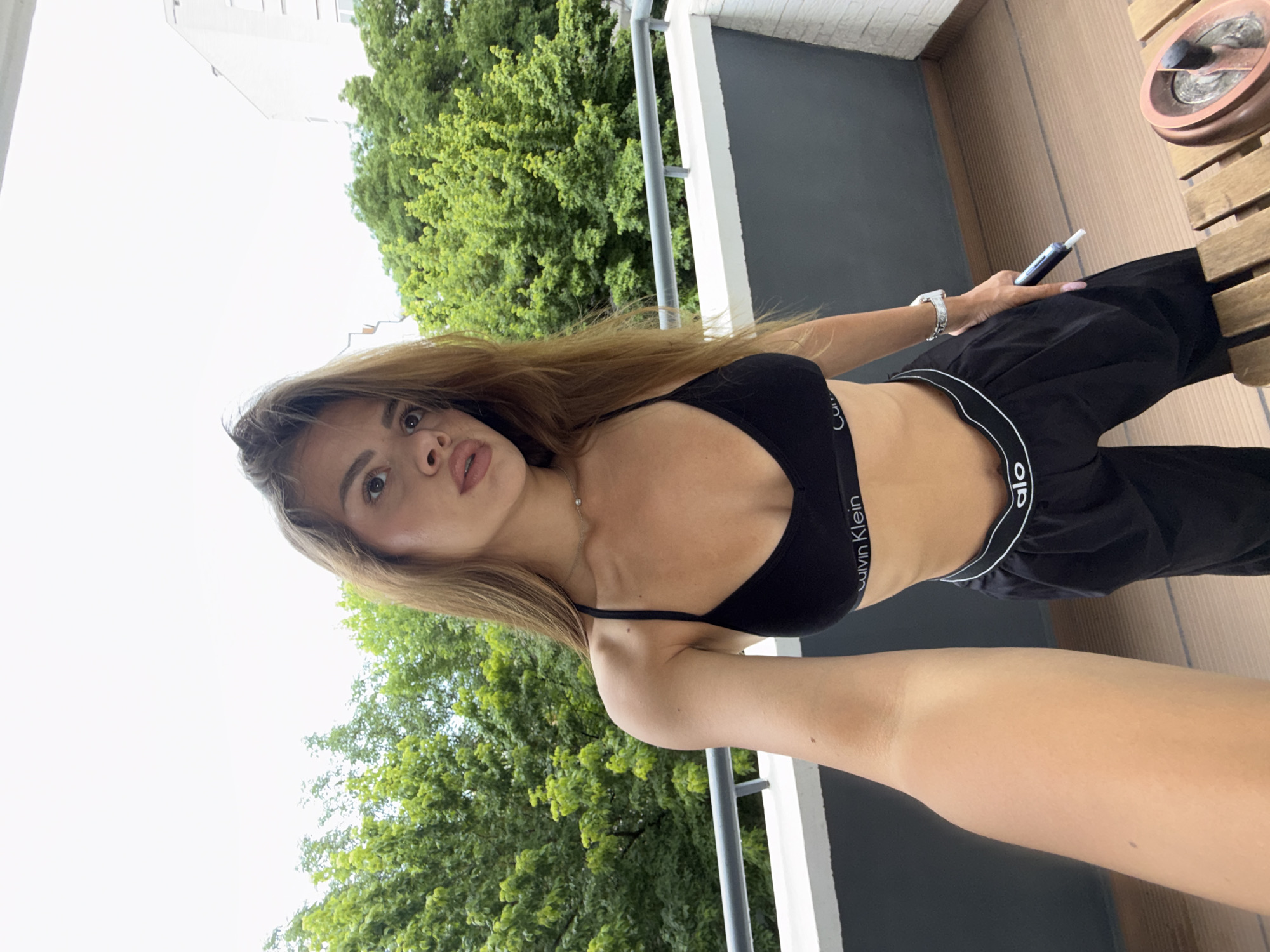 A young woman is captured in a selfie-style photograph, posing outdoors on what appears to be a balcony or terrace. She has long, light brown hair and is looking directly at the camera with a neutral expression and slightly parted lips. She is wearing a black Calvin Klein sports bra and black track pants with a visible "alo" branded waistband. A silver-toned watch adorns her left wrist, and she holds a black and blue electronic device (possibly a vape pen) in her right hand, which has long, pink-painted fingernails. A delicate necklace with a small pendant is visible around her neck.
The background features lush green trees and a bright, overcast sky, suggesting daytime. A light-colored building is partially visible in the upper background. To her right, a wooden slatted surface, likely a table or flooring, is visible, along with a circular, reddish-brown ashtray or dish. To her left, a dark grey wall or railing with a white support structure is present, reinforcing the balcony setting.