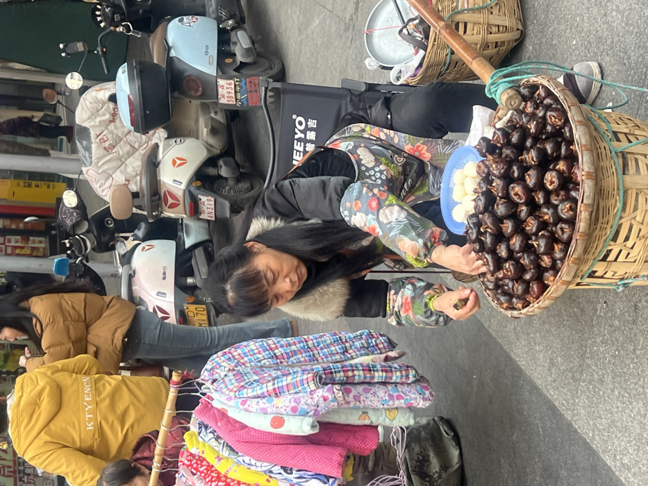 The image depicts an outdoor street scene in Jiangjin, Chongqing Municipality, China. A woman, centrally positioned and squatting or kneeling, is focused on a large bamboo basket filled with dark, cooked water chestnuts. She wears a black jacket with a light fur collar and a floral-patterned apron. Her left hand holds a single water chestnut, while her right hand is within the basket. Adjacent to her on the ground, a small blue bowl contains several white, peeled spherical items. To her right, another bamboo basket holds a metal pan and other indistinct items, with a segment of a wooden carrying pole visible. To her left, a portable clothing rack, constructed from a bamboo pole, displays a collection of colorful children's jackets and shirts. In the background, two individuals are partially visible: one wearing a yellow hooded jacket with "KTYENCN" printed on the back, and another in a brown puffer jacket and jeans. Behind these individuals, three electric scooters are parked, showing Chongqing license plates starting with "渝 D". The scooters vary in color (light pink/white, white/beige, light blue) and feature a quilted seat cover on one and a black storage box on another. The ground is paved, and blurred storefronts are visible in the far background.