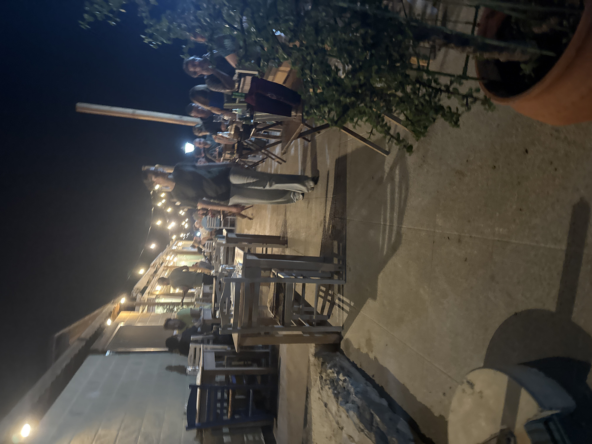 The image depicts an outdoor dining area at night, likely a restaurant or cafe, located in the Agios Tychona Community, Cyprus. The scene is illuminated by string lights and overhead lamps, casting a warm glow on the tables and patrons. Several tables, mostly rectangular and made of light-colored wood, are set with chairs and glassware. In the foreground, a person is walking away from the camera, dressed in a dark top and light-colored pants. Other individuals are seated at tables in the background, engaged in conversation or dining. A potted plant with green foliage is visible in the right foreground. The environment appears to be paved, with a stone border along the left side of the walkway. The building to the left has a light-colored facade and a covered awning. The overall atmosphere is relaxed and social, consistent with an evening dining experience.