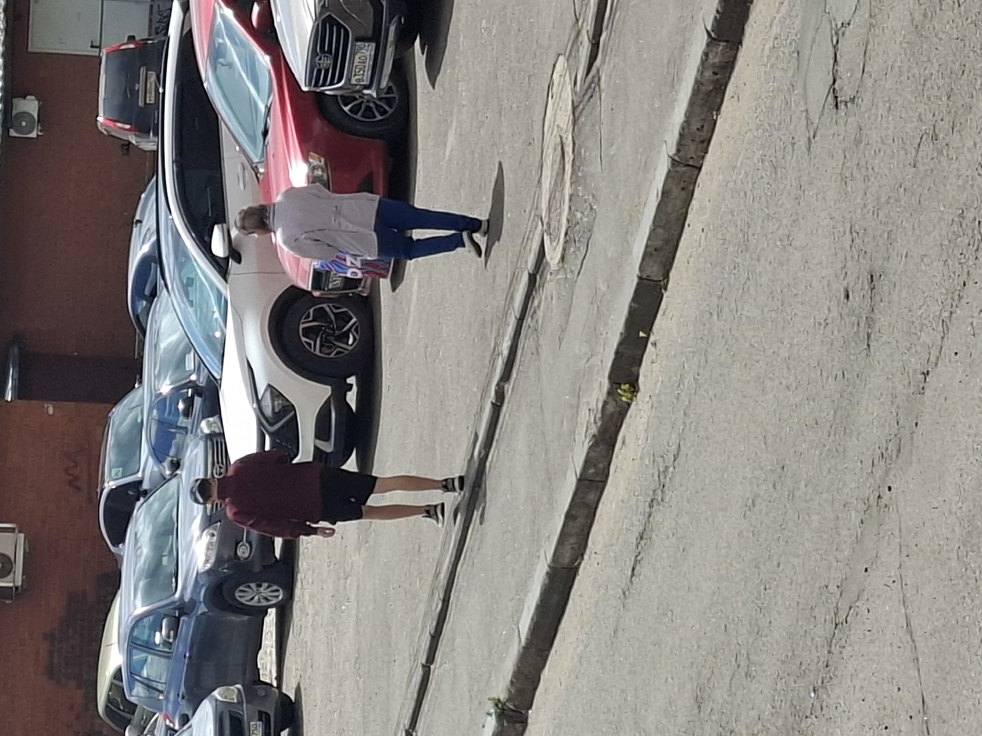 This image shows a street scene with several parked cars and two people. The first person is a woman wearing a light-colored jacket and dark pants, with blonde hair. She is standing next to a white car and appears to be walking or looking at something. The second person is a man wearing a dark maroon jacket and dark shorts, with dark hair. He is standing near a dark-colored SUV and appears to be walking or moving.
The scene is an outdoor urban environment, likely a parking lot or street, with a brick building visible on the left side and a paved surface with a curb and some greenery on the right. Several cars are parked in a row, including a red car, a white car, a dark-colored SUV, and other vehicles. The lighting suggests it is daytime, possibly sunny given the shadows cast by the people and cars. The overall impression is a casual, everyday scene. There is no visible text in the image.