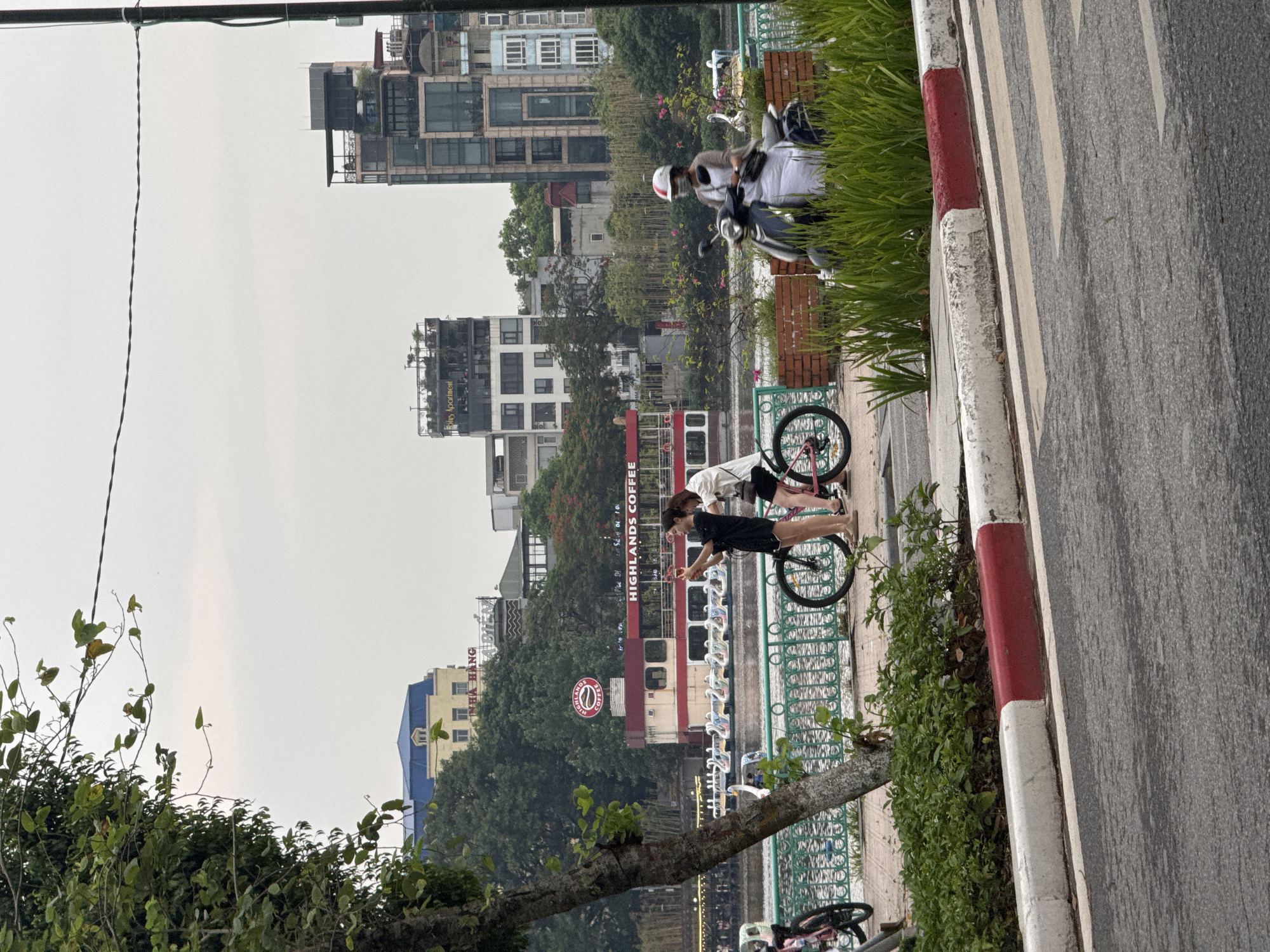 This image depicts a street scene in Hanoi, Vietnam, likely near a lake or body of water. In the foreground, a paved road with white and red curbs is visible. A sidewalk with green bushes and a red and white striped curb separates the road from a railing overlooking a waterway. Beyond the railing, two young women are standing next to bicycles. One woman, wearing a black outfit, is taking a selfie with the other woman. The second woman is wearing a white shirt and black shorts. In the middle ground, a red and white boat is docked, with the "HIGHLANDS COFFEE" logo prominently displayed. Several swan-shaped paddle boats are lined up in the water. In the background, various buildings are visible, including a yellow building with a blue roof and modern, multi-story apartment buildings. A person wearing a helmet and a mask is riding a scooter on the right side of the frame. The sky is overcast, suggesting a cloudy day. The overall scene portrays a casual urban environment with elements of leisure and commerce.