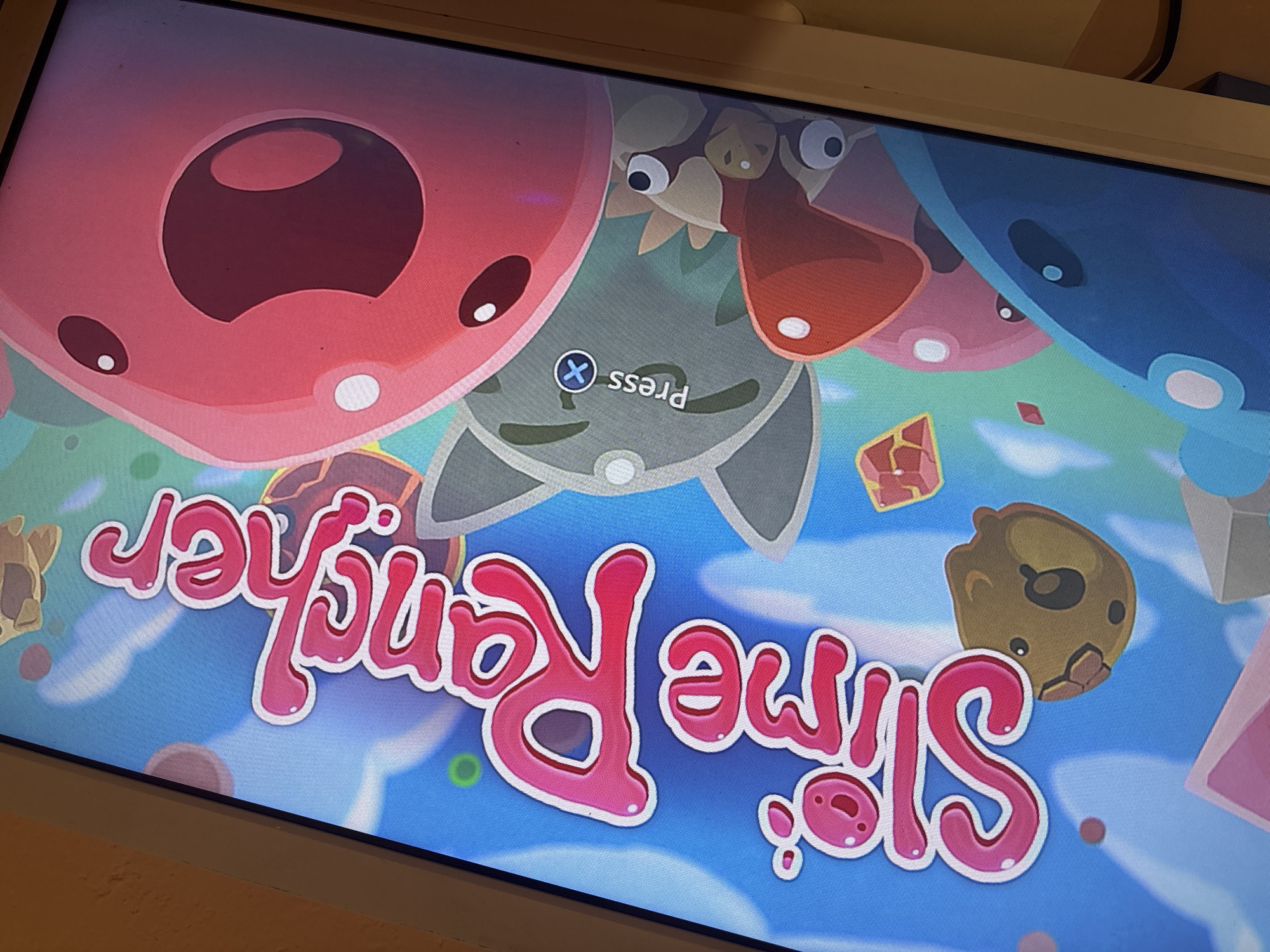 The image is a close-up shot of a television screen displaying the title screen of the video game "Slime Rancher". The screen features a colorful background with various cartoonish slime creatures of different colors (pink, gray, brown, blue) arranged across the top of the screen. In the middle of the screen, the game's title "Slime Rancher" is written in large, stylized pink lettering. Above the title, the prompt "Press X" is visible, indicating a control input on a console. The background includes blue sky elements and various geometric shapes.
The image was not taken in Lom, Bulgaria. It appears to be in a private residence.