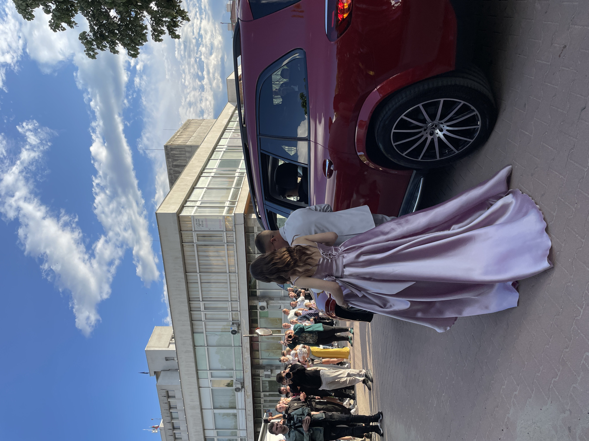 A young woman in a floor-length, light pink satin gown is being assisted from the back seat of a maroon SUV. A male figure is visible, leaning into the vehicle. A crowd of people, some holding cameras, stands along the side of a building. The building is a multi-story structure with a grid of windows. Above the building are clouds and blue sky. The ground is paved with light red bricks. The building is the "Zlatograd Municipality", in Zlatograd, Bulgaria.