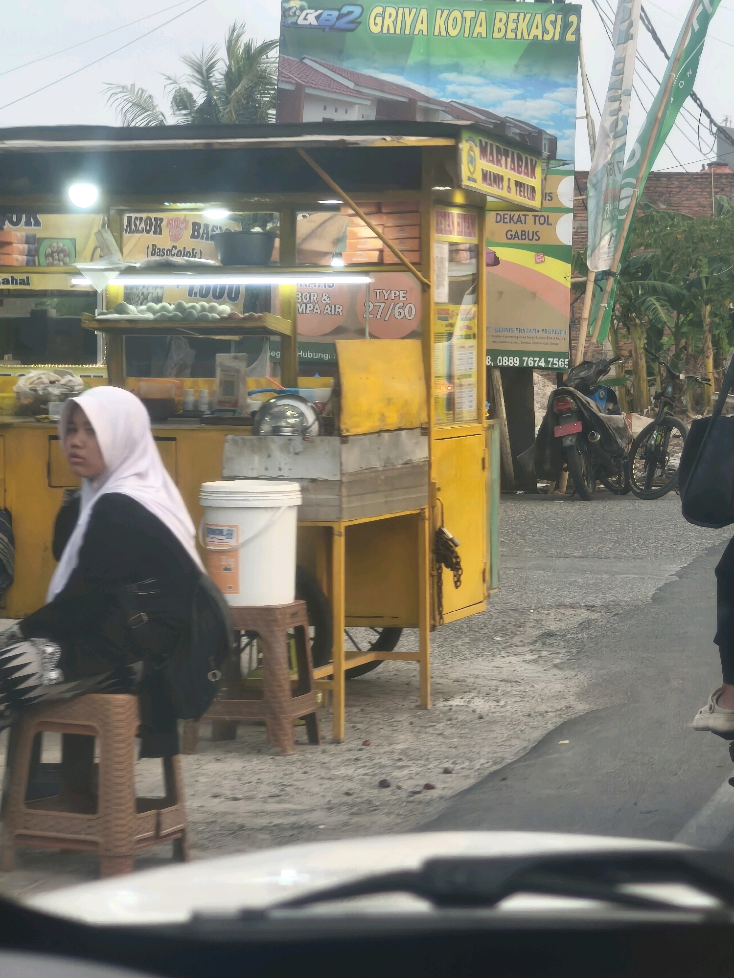 The image depicts a street scene in Satriamekar, Indonesia, featuring a food stall with a prominent yellow facade. The stall displays signs advertising "MARTABAK MANIS & TELUR" (sweet and egg martabak) and "ASLOK BASLOK (BasoColok)". Above the stall, a large green banner reads "GRIYA KOTA BEKASI 2". In the foreground, a person wearing a white hijab and a dark top is seated on a brown stool. Behind the stall, two motorcycles are parked, one covered with a dark cloth. The ground is paved with asphalt and gravel. In the upper portion of the image, palm fronds are visible against a cloudy sky.